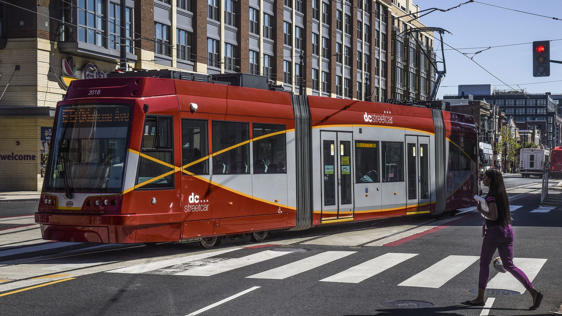 A red and gray dc streetcar with yellow diagonal accents travels along tracks in a city street; a woman in purple walks across a zebra crosswalk as buildings and overhead wires loom above.