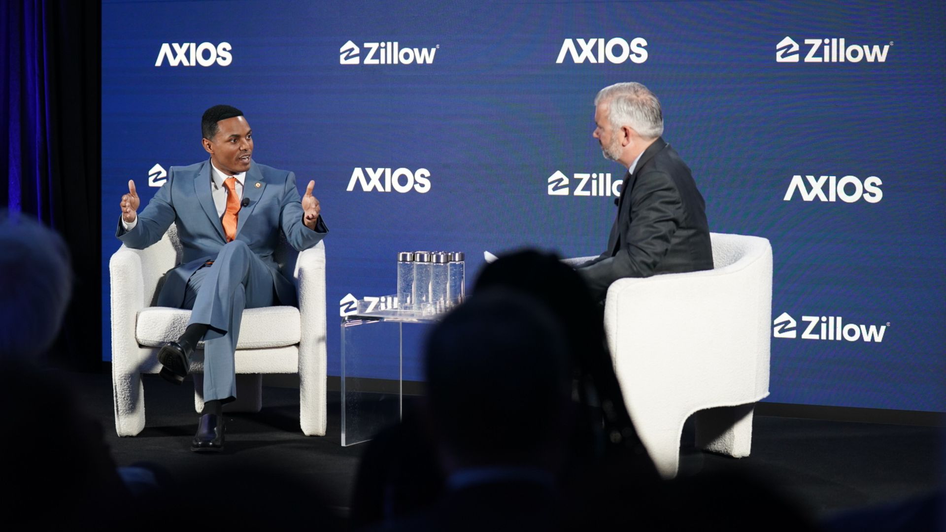 Two men in suits sit on white chairs on a stage with a blue backdrop showing Axios and Zillow logos; the left speaker gestures with an orange tie while the right listens, foregrounded by silhouettes.