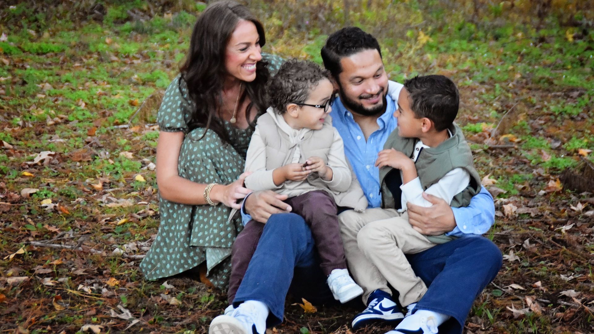 Family of four sits on autumn leaves in a park, smiling and cuddling: mother in green dress, father in blue shirt, and two kids in jackets enjoying time together.