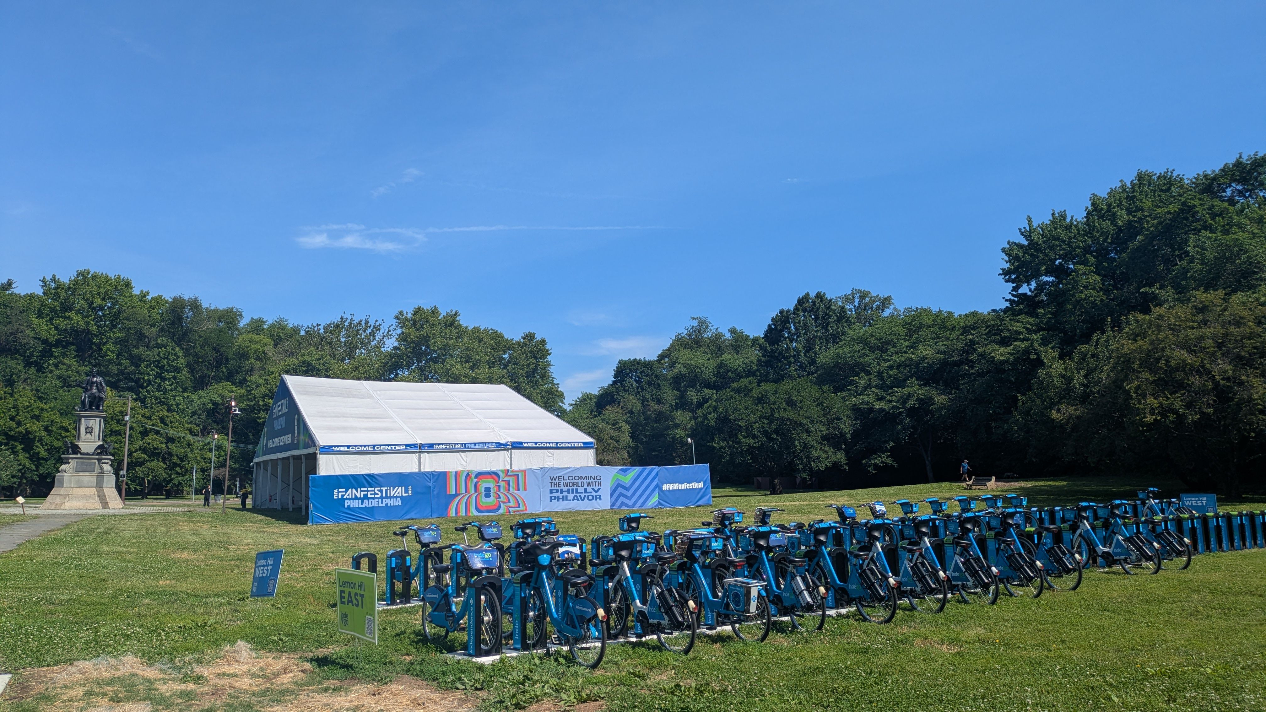 Bright outdoor scene with a row of blue rental bikes on a grassy field, a white event tent with "FAN FESTIVAL PHILADELPHIA" in front, a statue to the left, and green trees under a blue sky.
