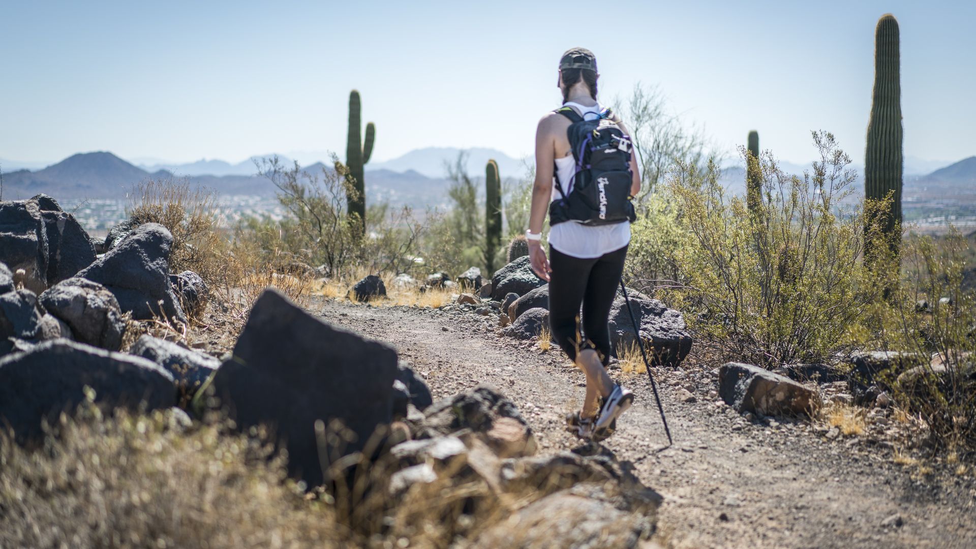 A woman hikes on a mountain trail in the desert surrounded by rocks and saguaro cactus. 