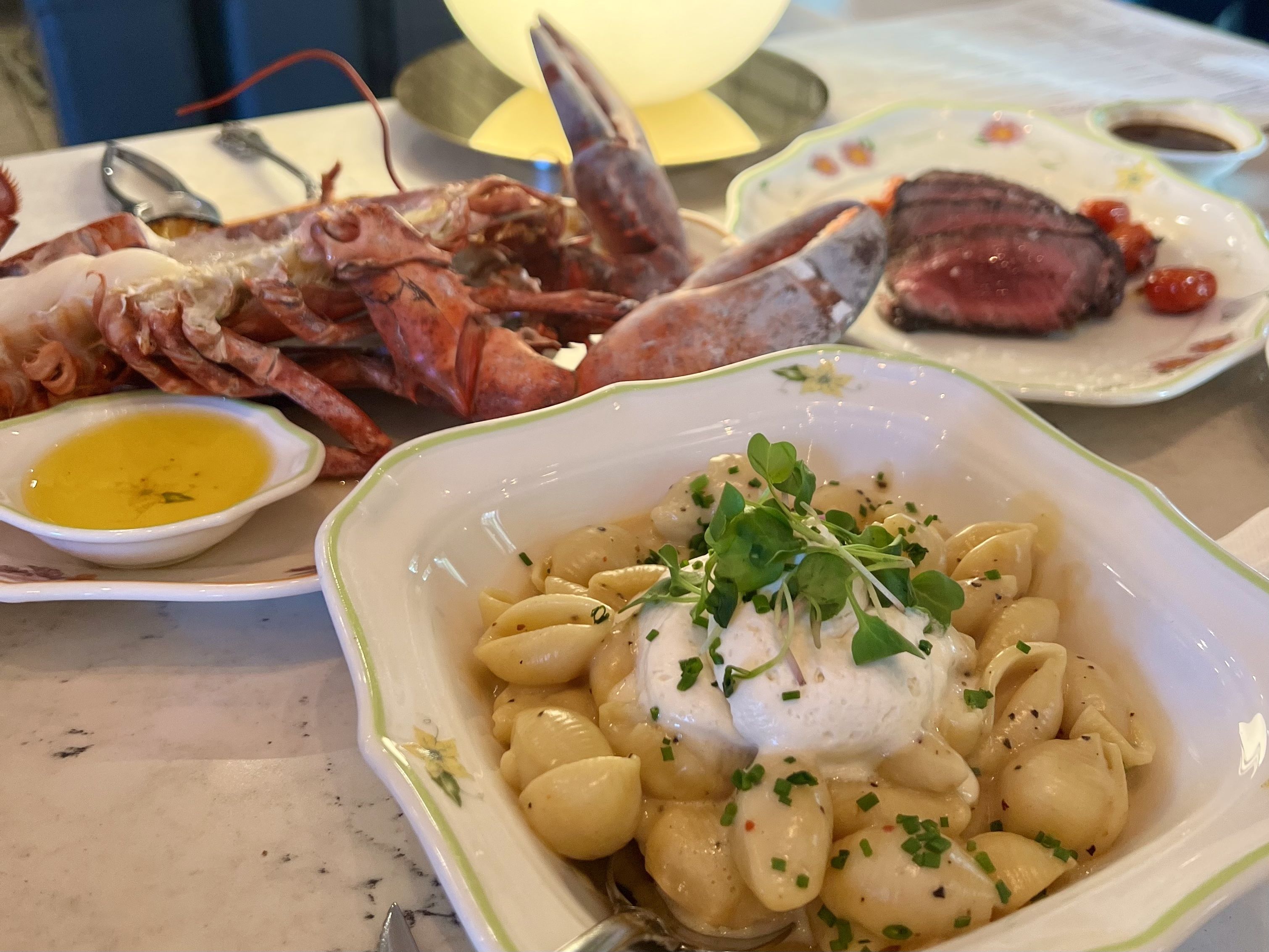 Platter shows lobster tails and shell pieces on the left, a seared pink-centered steak on a floral plate to the right, and creamy pasta with herbs in the foreground, plus yellow sauce on the side.