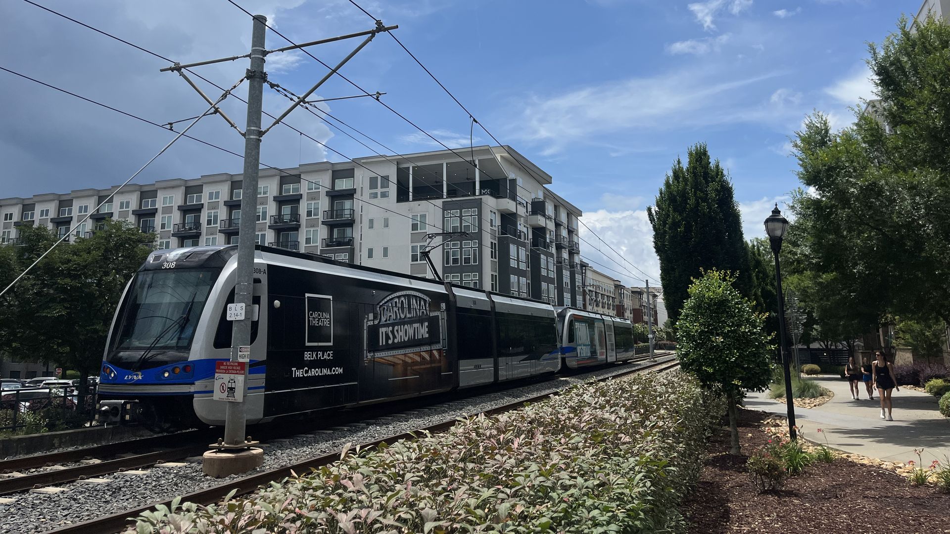 A modern tram on elevated tracks with overhead wires passes a contemporary apartment building; blue sky, white clouds, green trees, and pedestrians on a sunny sidewalk.