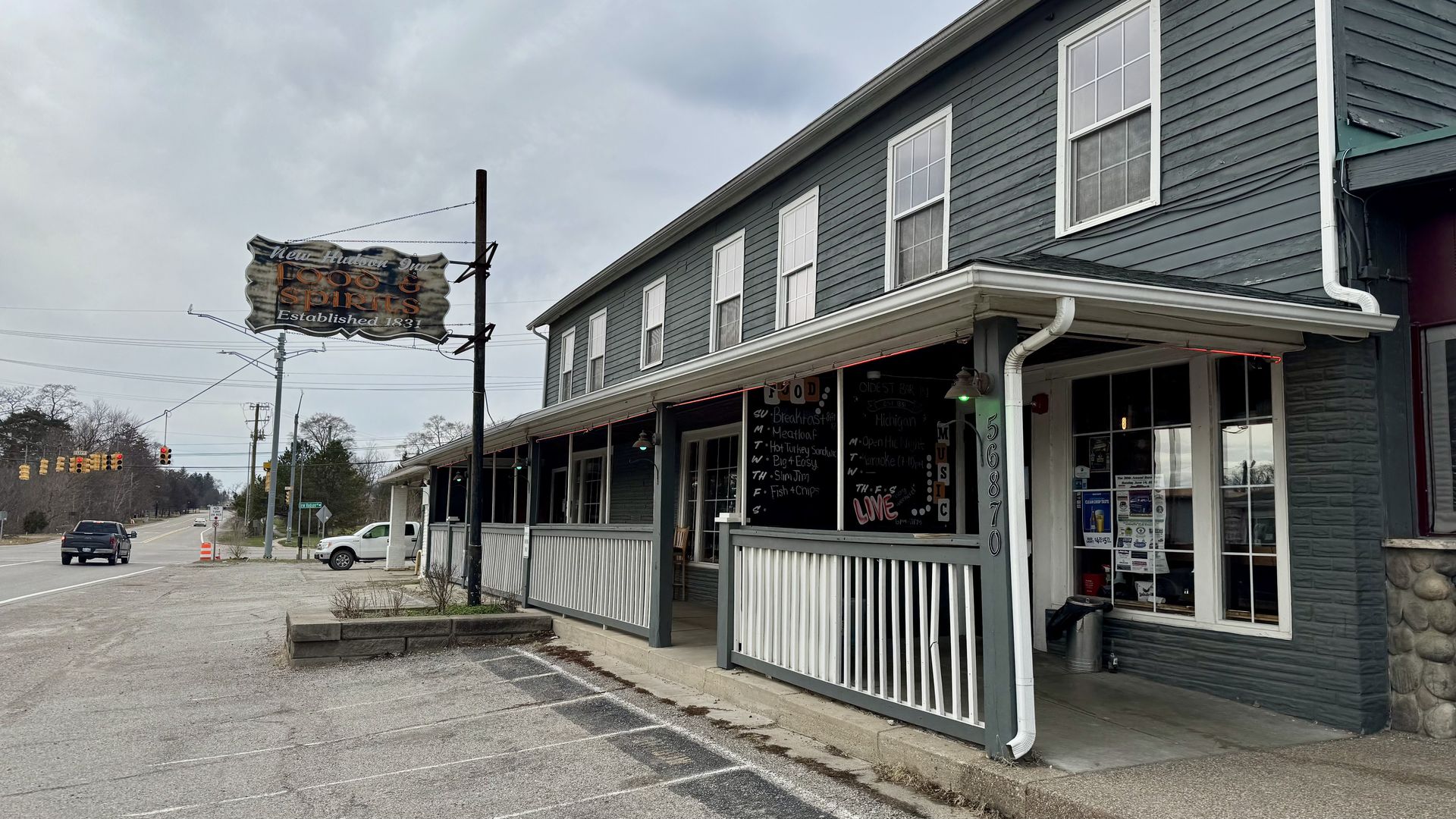Gray-blue two-story building with a long porch and white railings on a street; a weathered sign and chalkboard menus on the side of the storefront.
