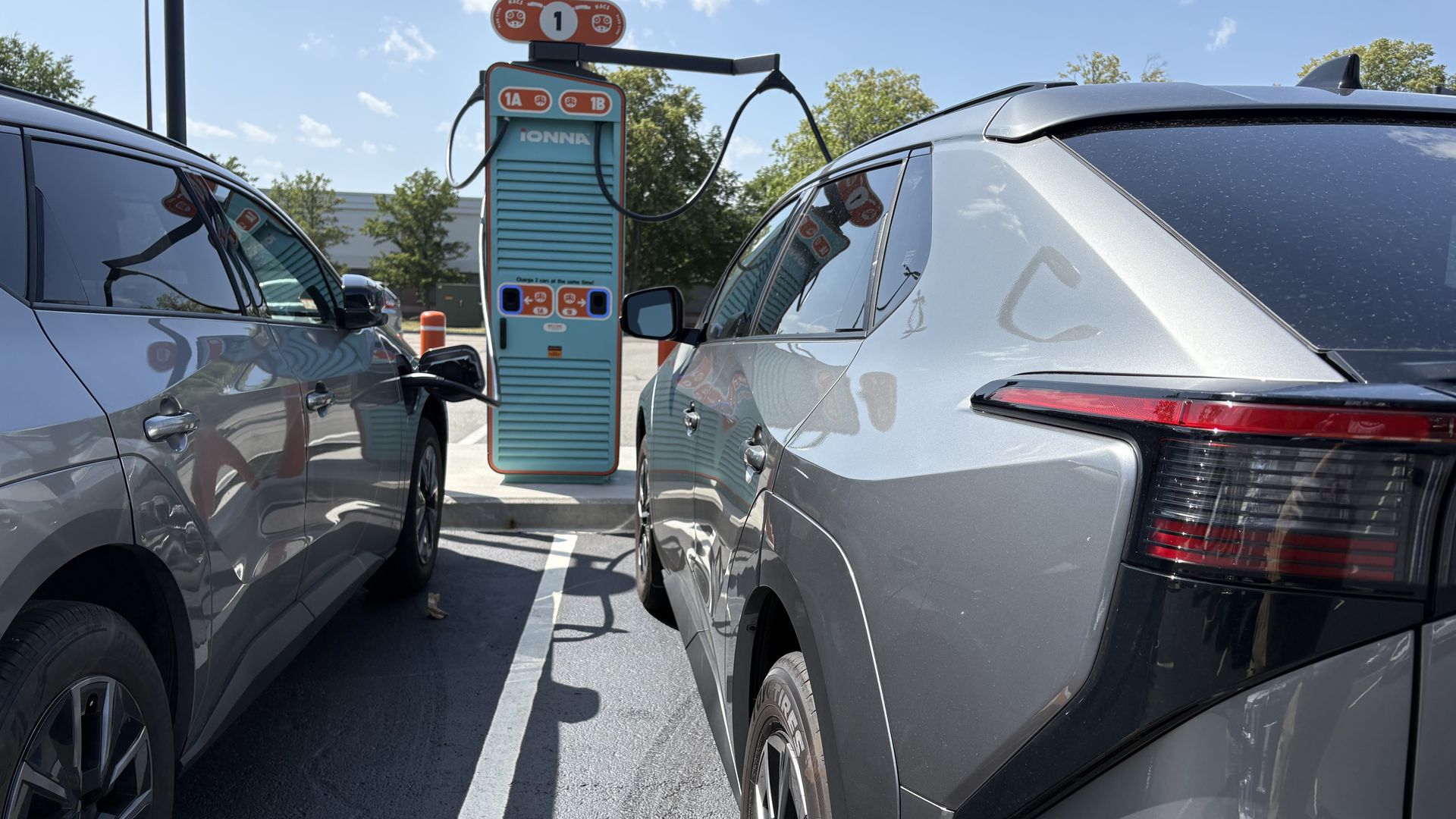 Two silver SUVs flank a teal retro-style Ionna EV charging station outdoors; cables connect to the cars as a bright blue sky and trees loom in the background.