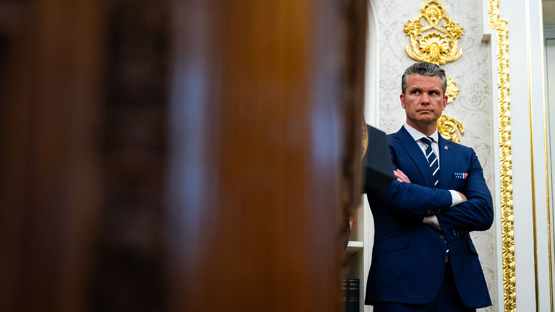 Pete Hegseth, a man, stands with his arms crossed. He is wearing a blue suit and blue tie with white-ish stripes. There are golden flourishes behind him on the wall.