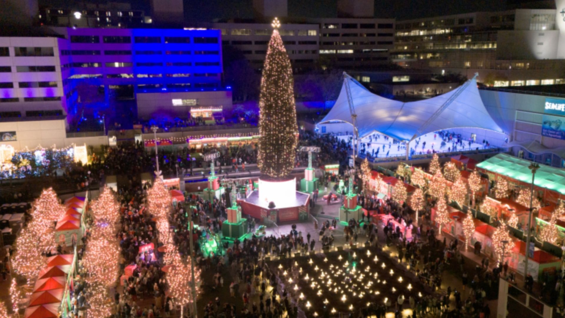 A bustling nighttime Christmas market with a large decorated tree center, lighted smaller trees, an ice skating rink under white canopy, and crowds amid festive red booths and buildings.