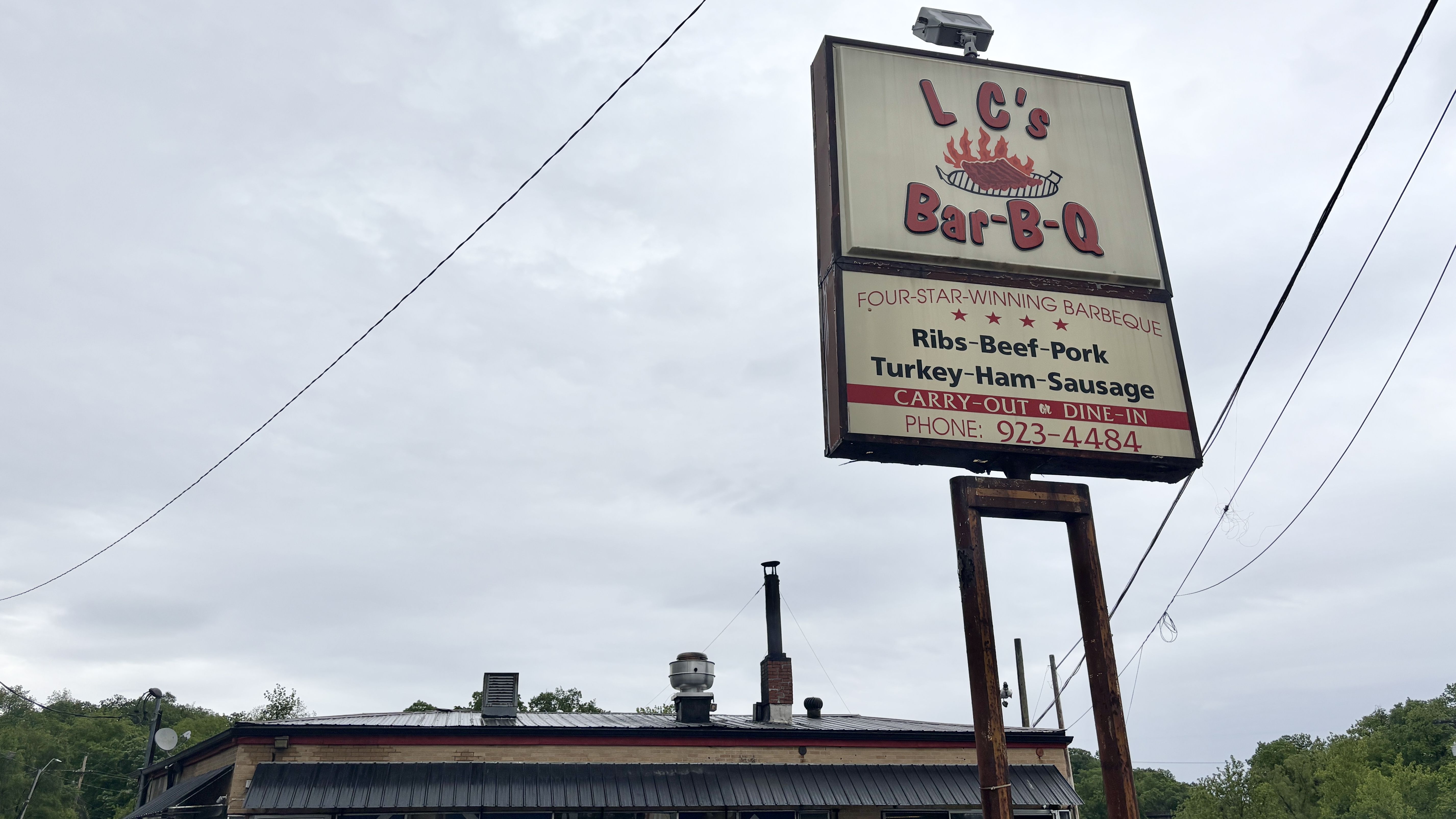 Tall outdoor sign for "LC's Bar-B-Q" with a flaming grill graphic. It lists ribs, beef, pork, turkey, ham and sausage; carry-out or dine-in, phone 923-4484, against an overcast sky.