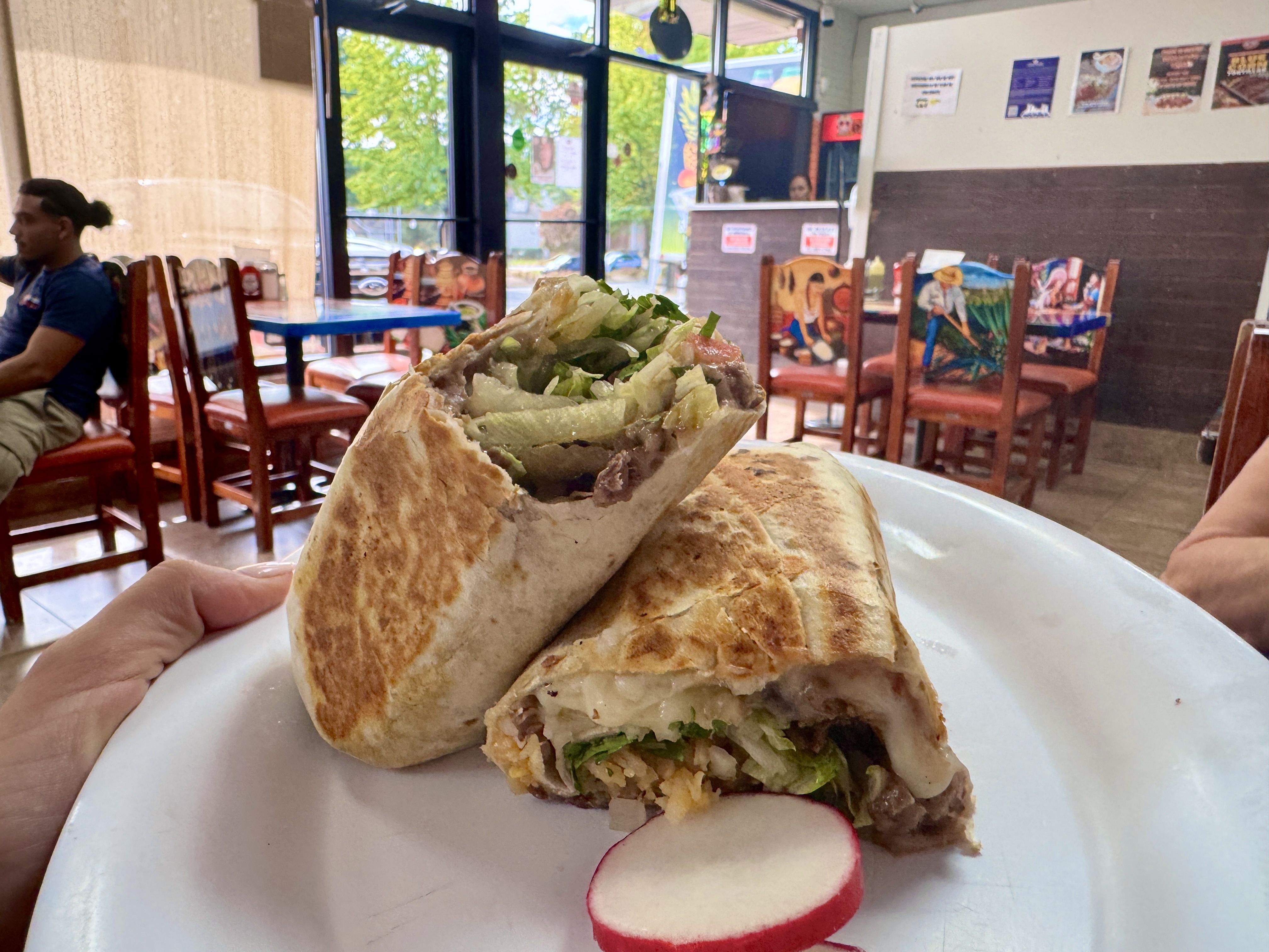 Close-up of a wrap filled with lettuce and veggies, held over a white plate. Inside a casual cafe with wooden chairs and glass doors; a man sits at a nearby table.