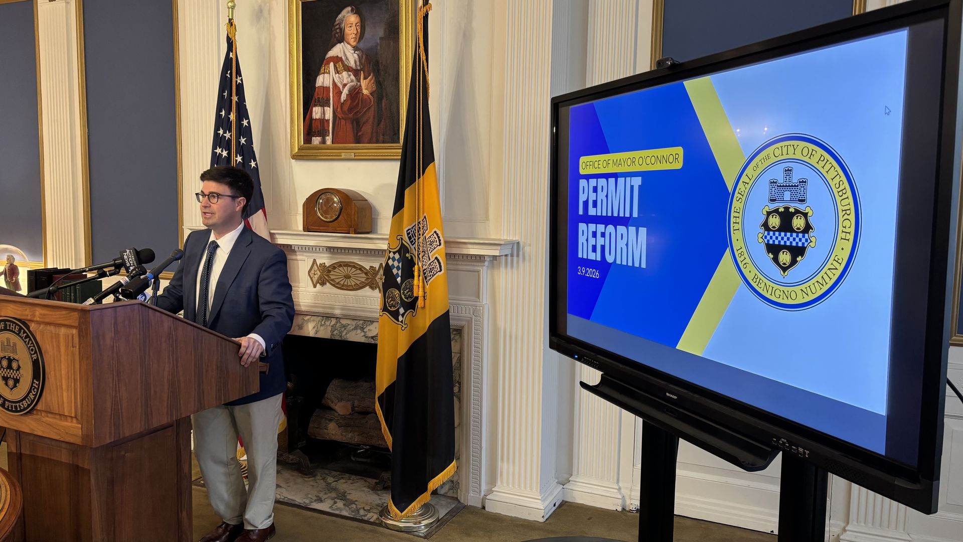 Man in a navy suit speaks at a wooden podium with microphones in a formal room, flanked by US flag and Pittsburgh city flag; a screen reads "PERMIT REFORM" with the city seal.
