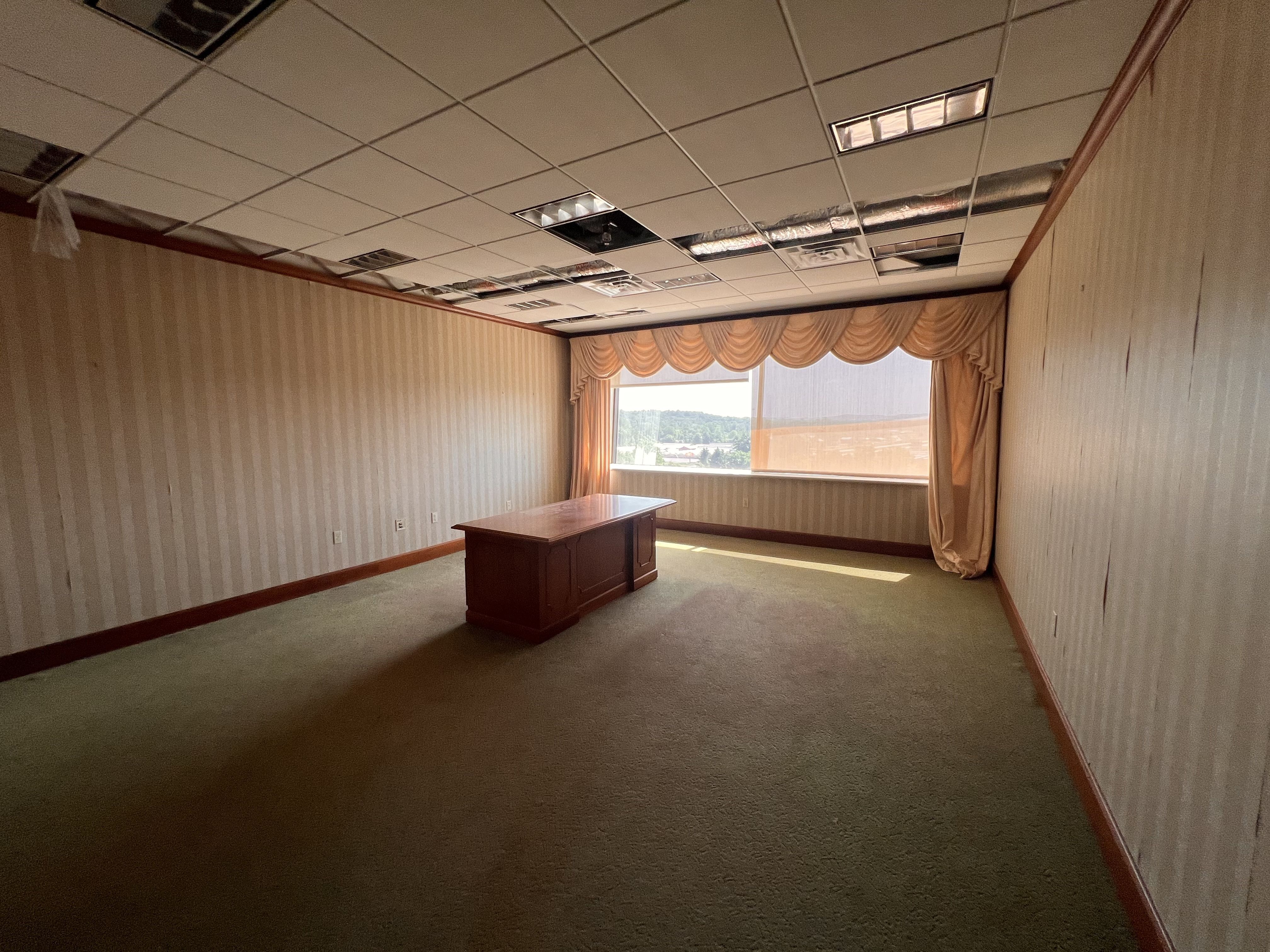An empty office/meeting room with beige striped walls, a single wooden desk in the center, and a window with peach drapes. The drop ceiling shows missing tiles and exposed ducts.