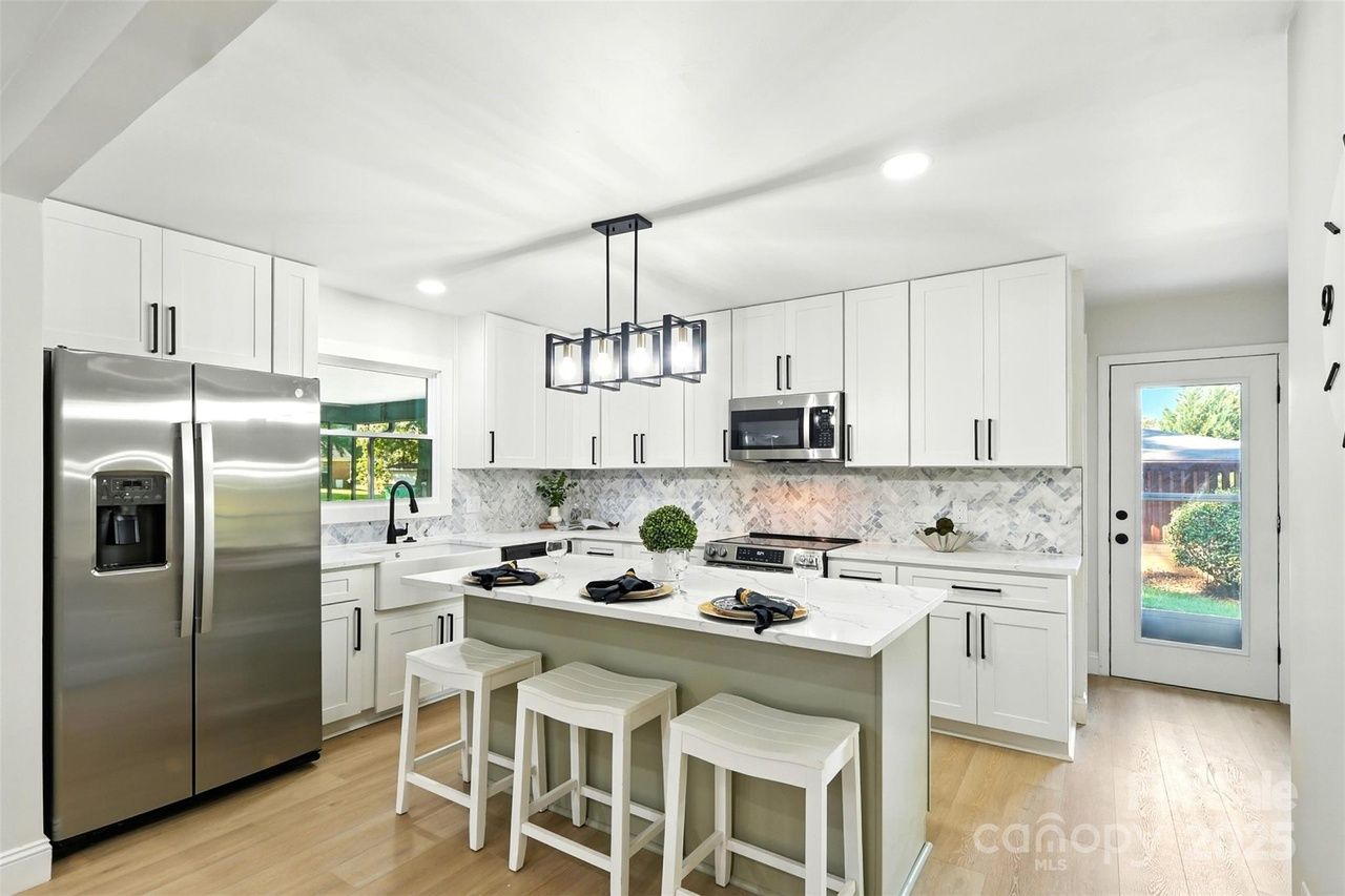 Modern all-white kitchen with marble backsplash, stainless steel fridge and microwave, island with three white stools, wood flooring, and a glass door leading outside.
