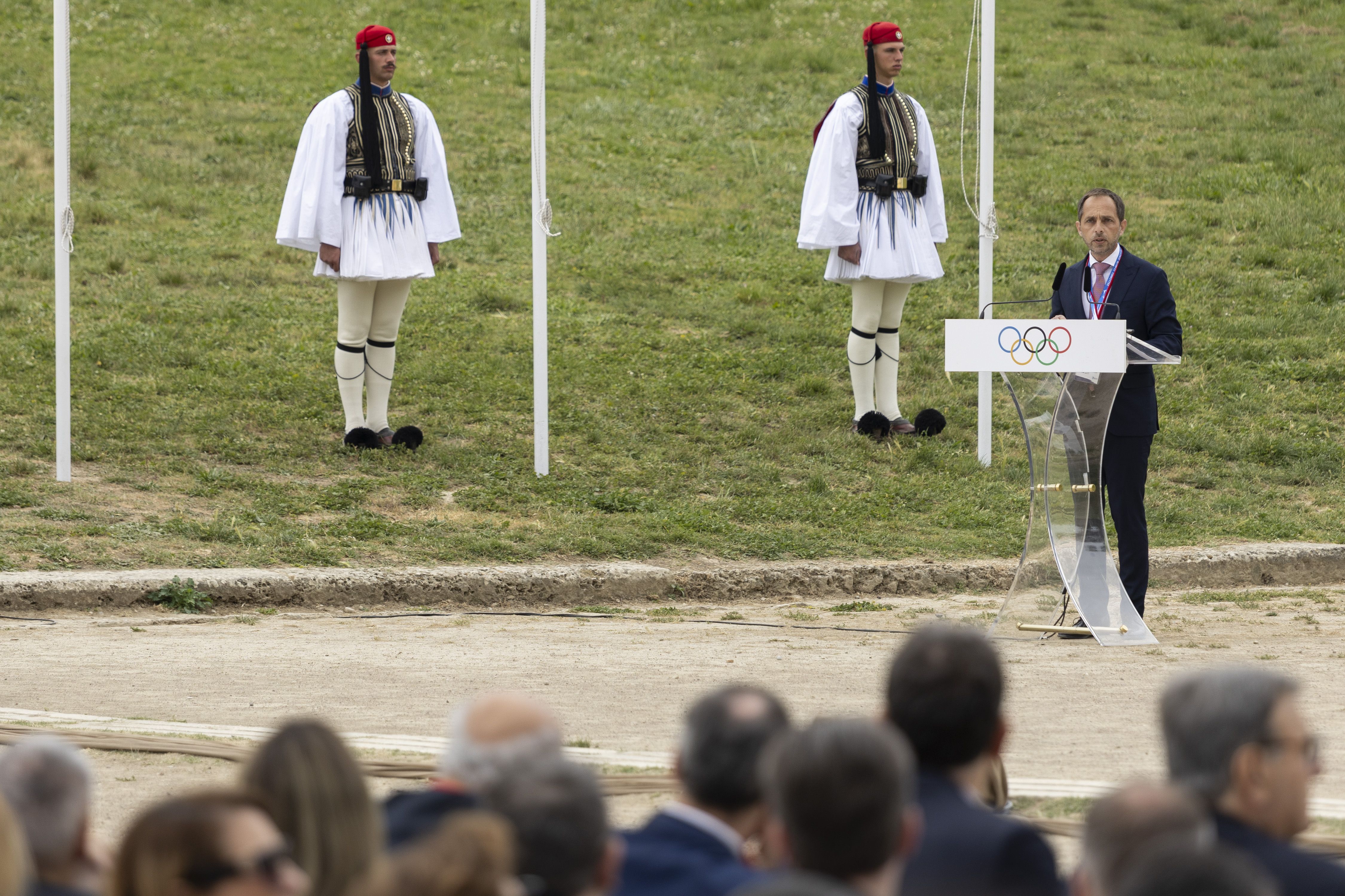 A man stands at a lectern giving a speech to a crowded audience outdoors. 