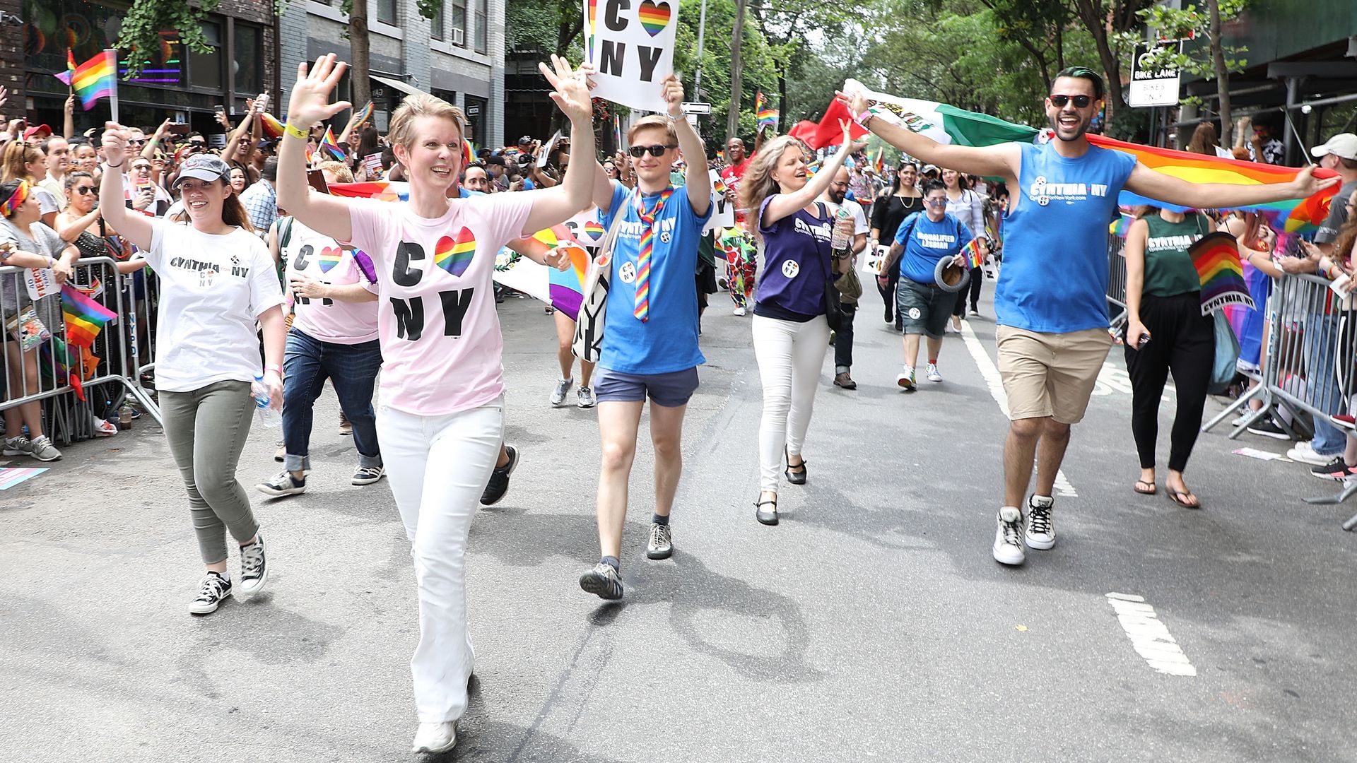 Colorful Pride parade on a city street with diverse marchers waving rainbow flags and smiling; a central participant wears a pink shirt and waves at the crowd.