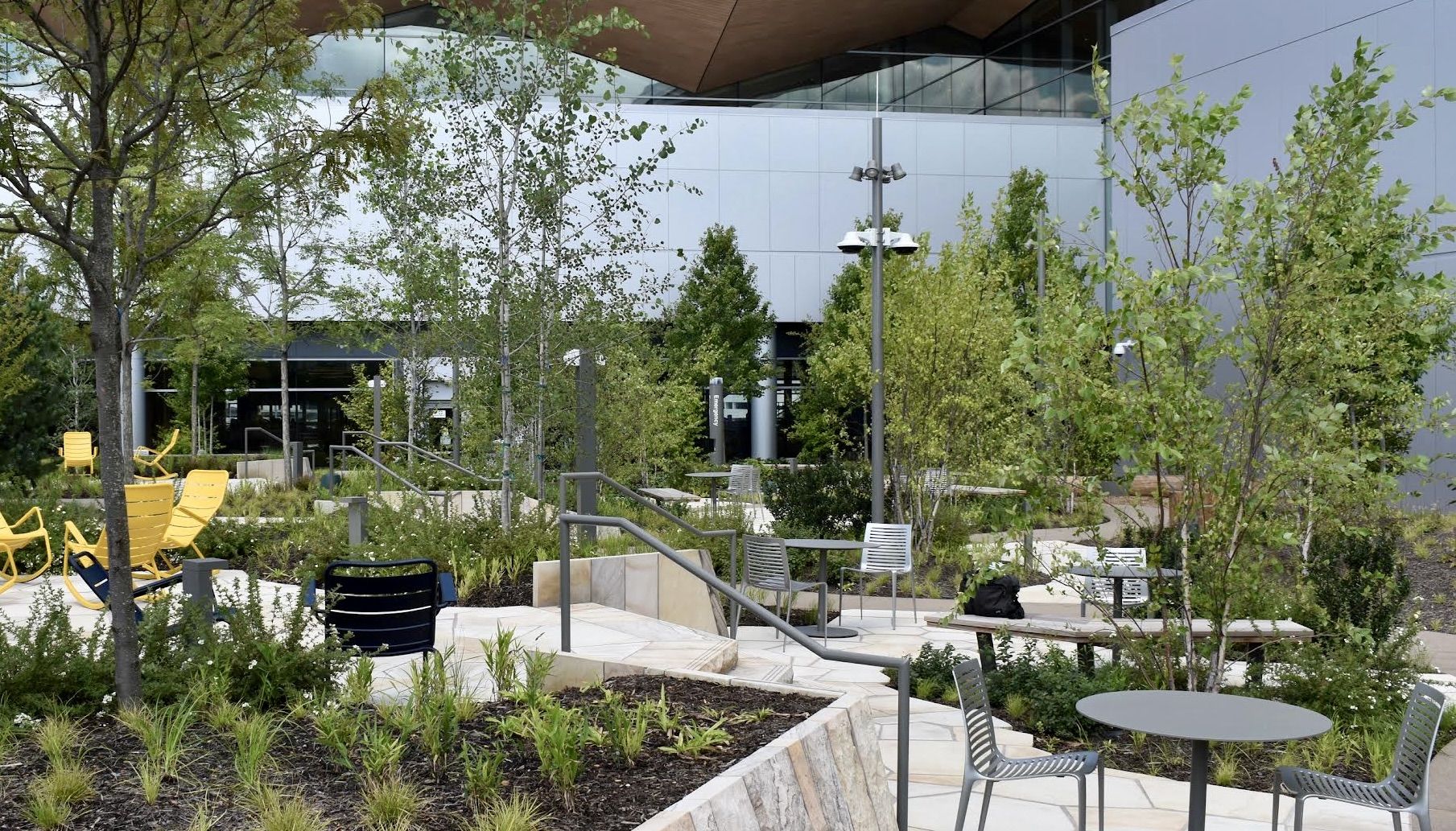 An outdoor courtyard beside a modern glass-building: gray metal chairs and round tables among green trees, with bright yellow lounge chairs to the left.