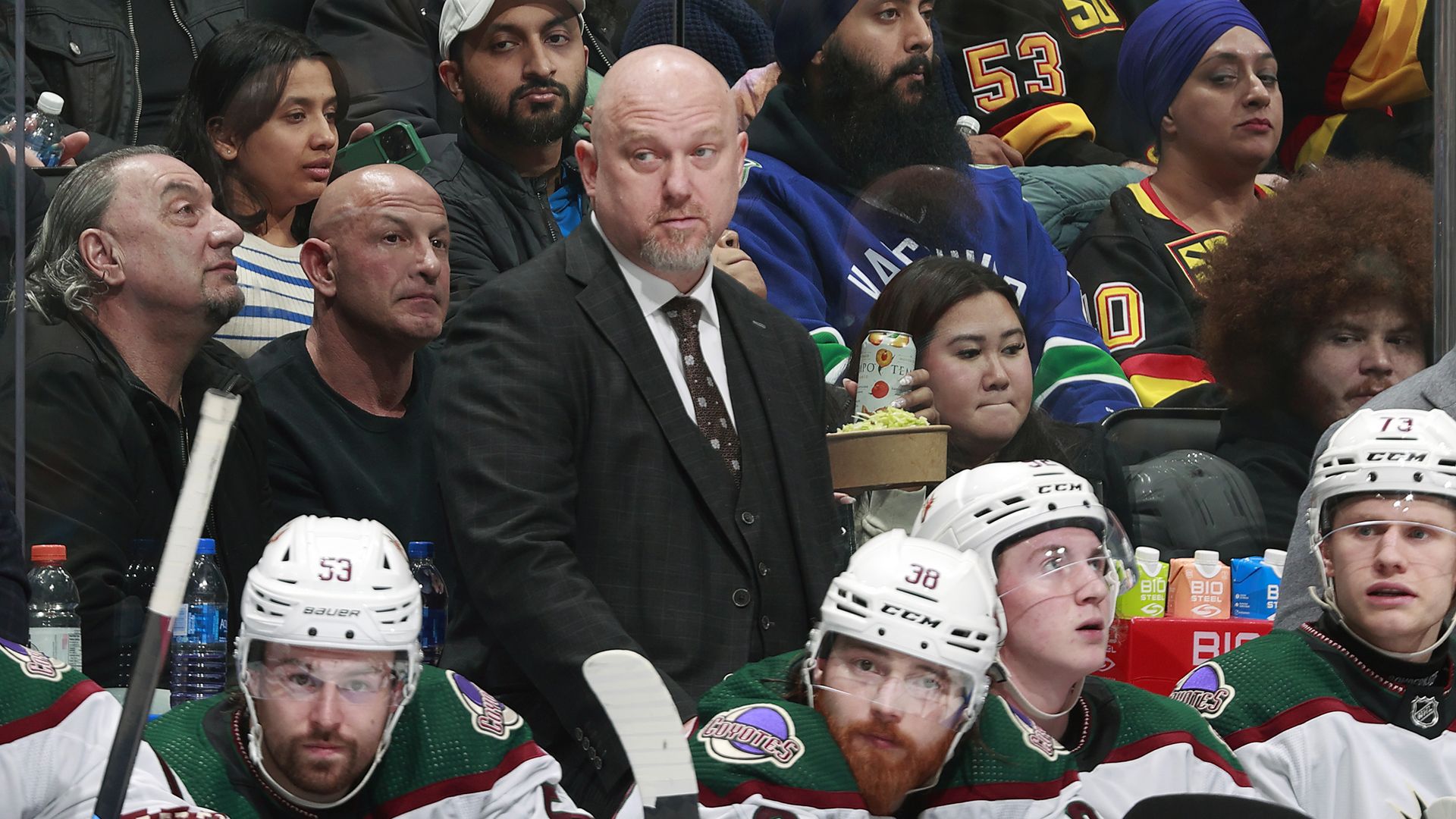 A hockey coach stands behind several players seated on the bench.