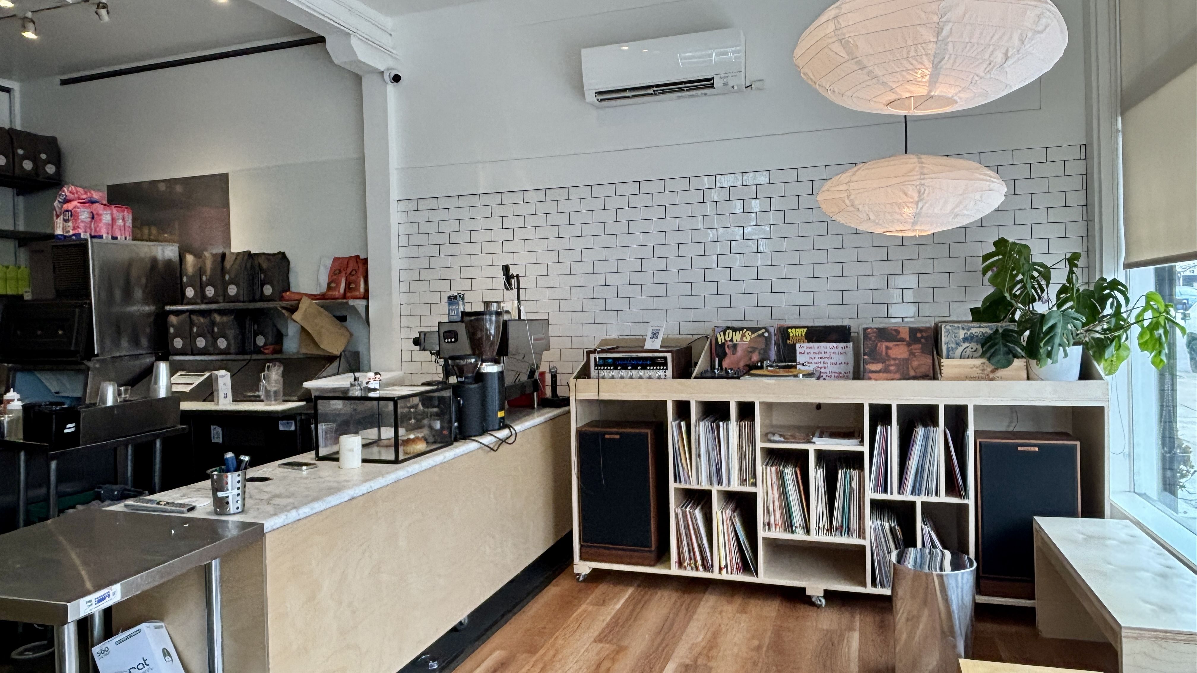 Bright cafe with a white subway-tiled wall, a counter with espresso gear, shelves of coffee bags and vinyl, two white paper lanterns overhead, and a large Monstera by the window.