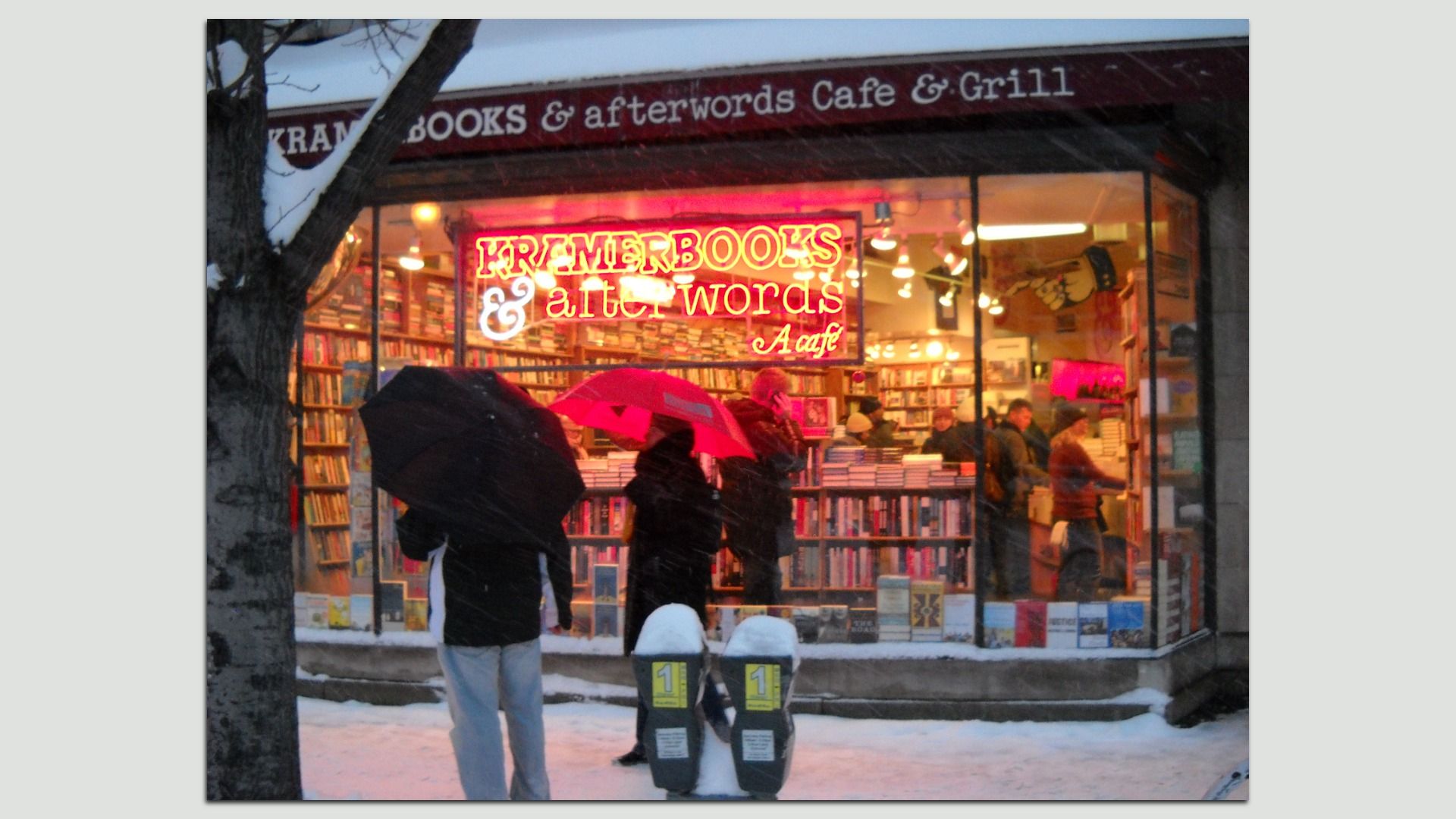 Snowy storefront of Kramer Books & Afterwords Café, with a neon red sign glowing in the window. People with red and black umbrellas stand outside while warm lights reveal shelves of books inside.