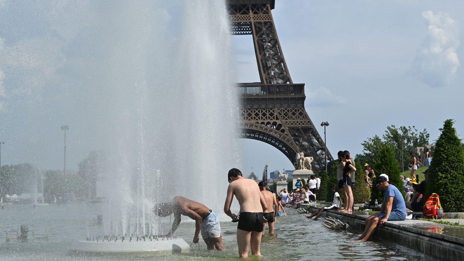 A wide fountain shoots water into a pool while people in swimwear stand along the edge; in the background is a brown Eiffel Tower replica under a blue sky.