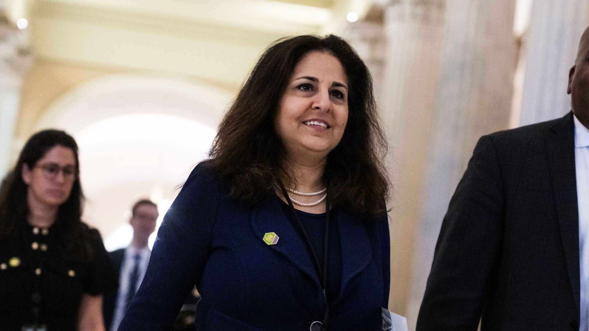 Neera Tanden, wearing a blue blazer, walks through a well-lit hallway with white marble columns.
