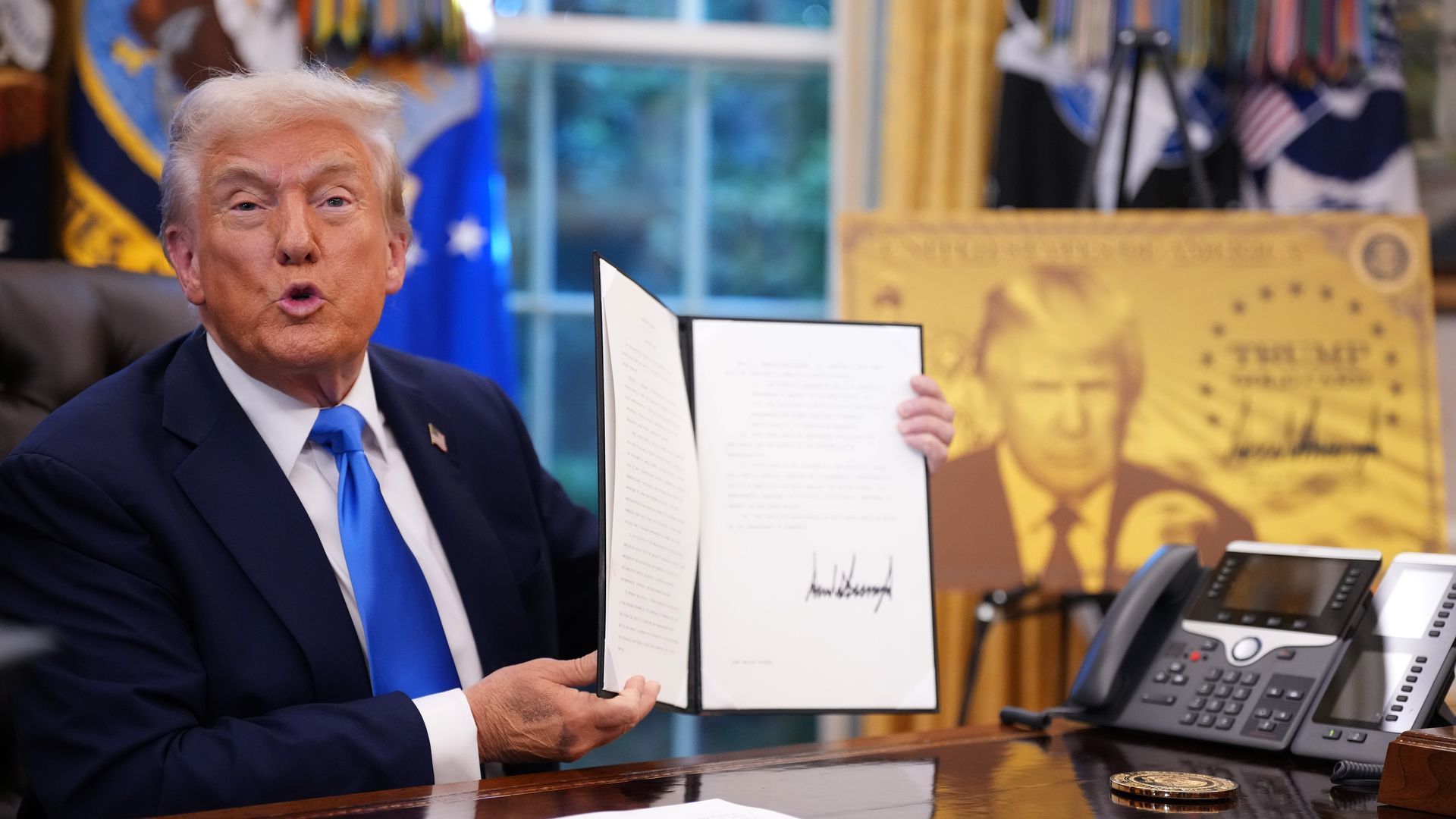 President Trump, seated at his desk and holding up an order bearing his signature. 