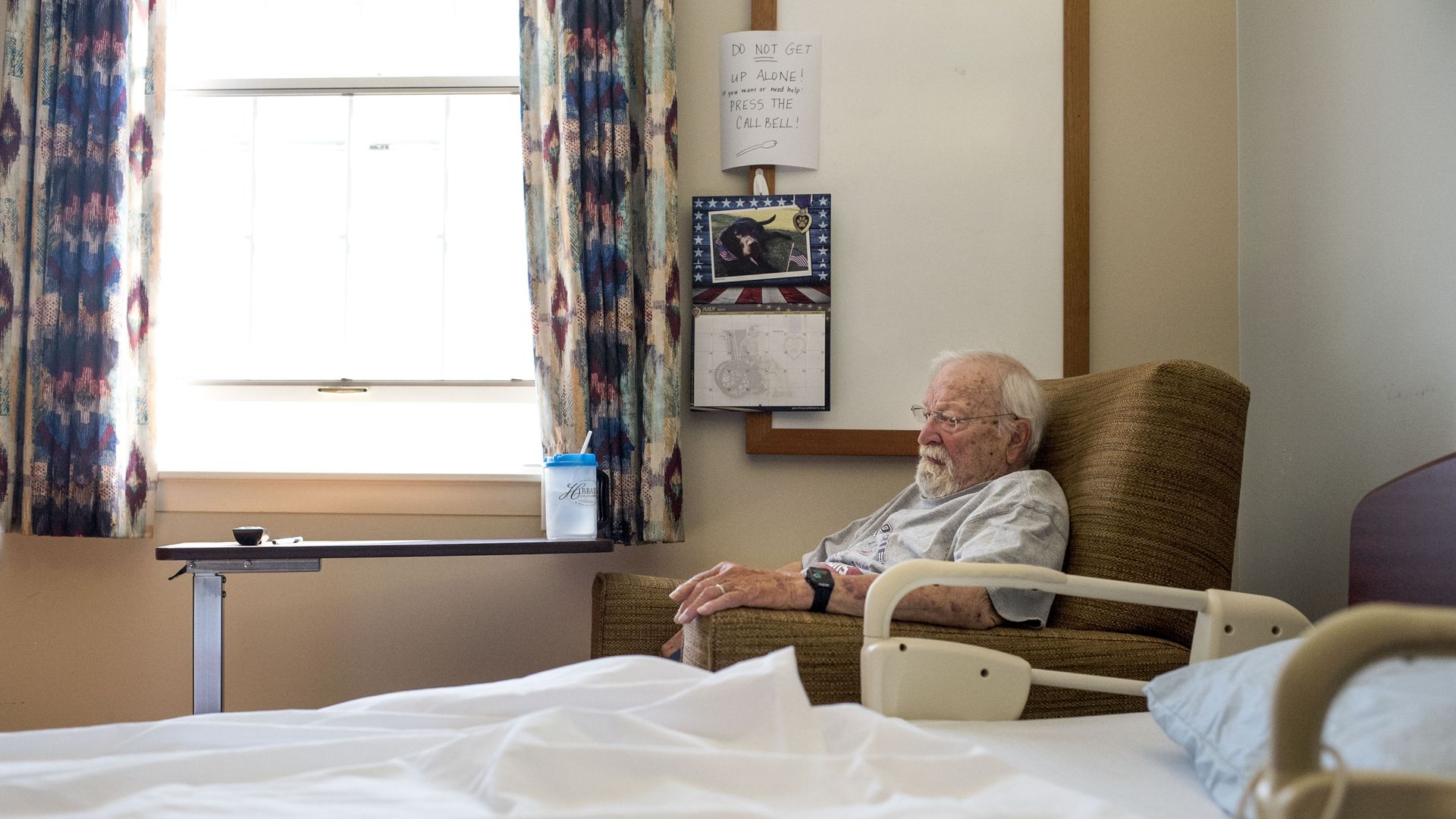 In this image, a man sits in an armchair in a nursing home room, near an open window that is brightly lit, and his bed.
