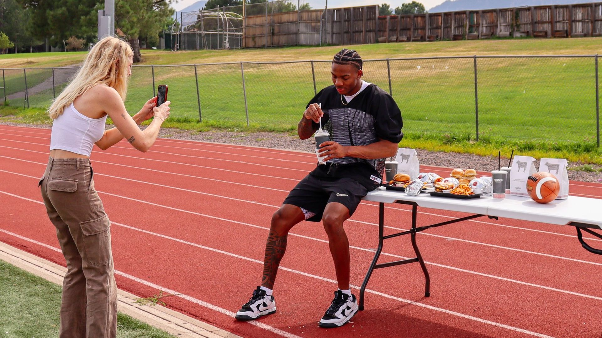 DJ McKinney, in black sportswear, sits on a table with fast food and a football on a track field, while a woman takes a video of him with her phone.