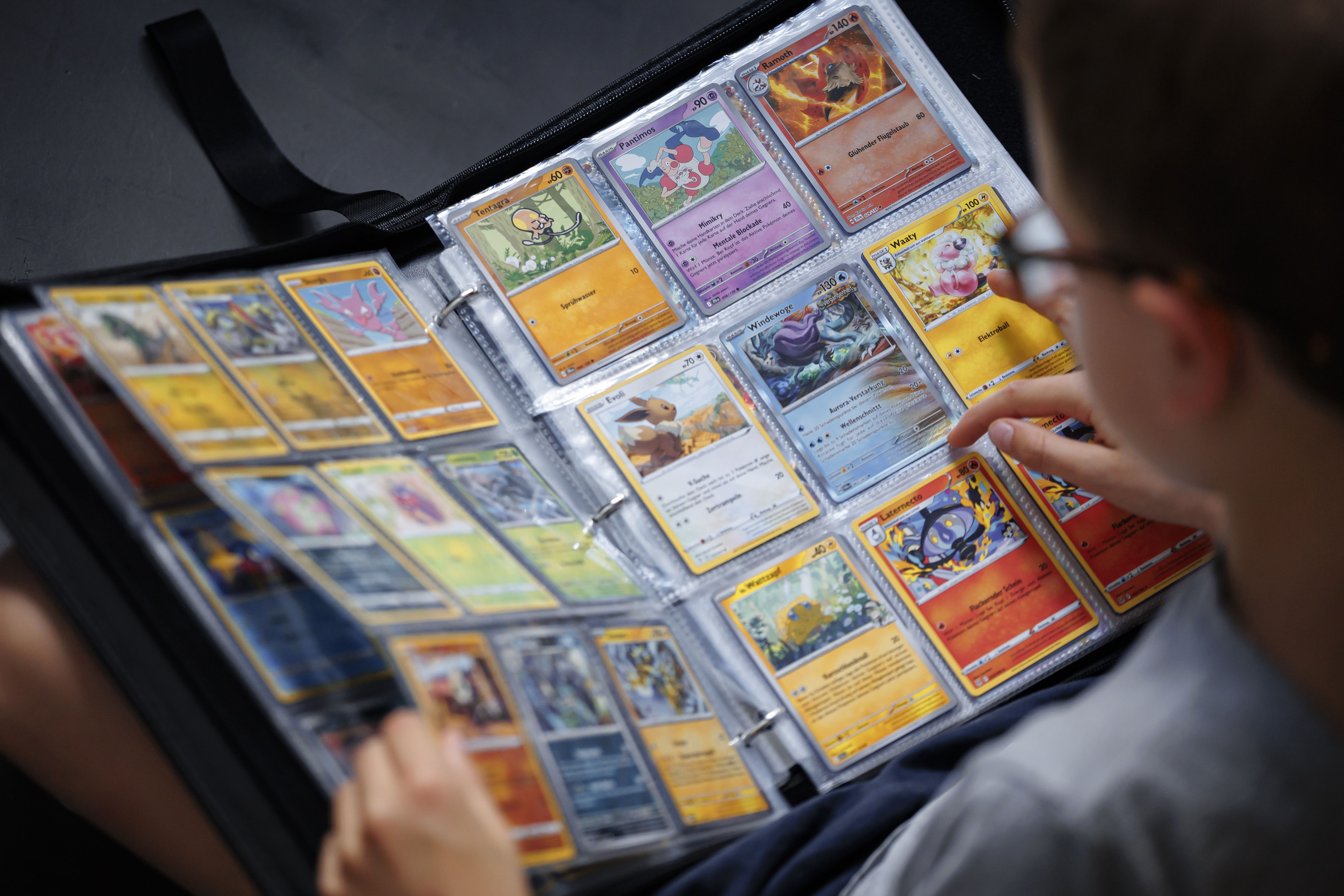 Person browsing a binder filled with colorful Pokémon cards, including Tentagra, Panfimos, Ramoth, Waaty, Eevee, and Laternecto, on a dark surface.