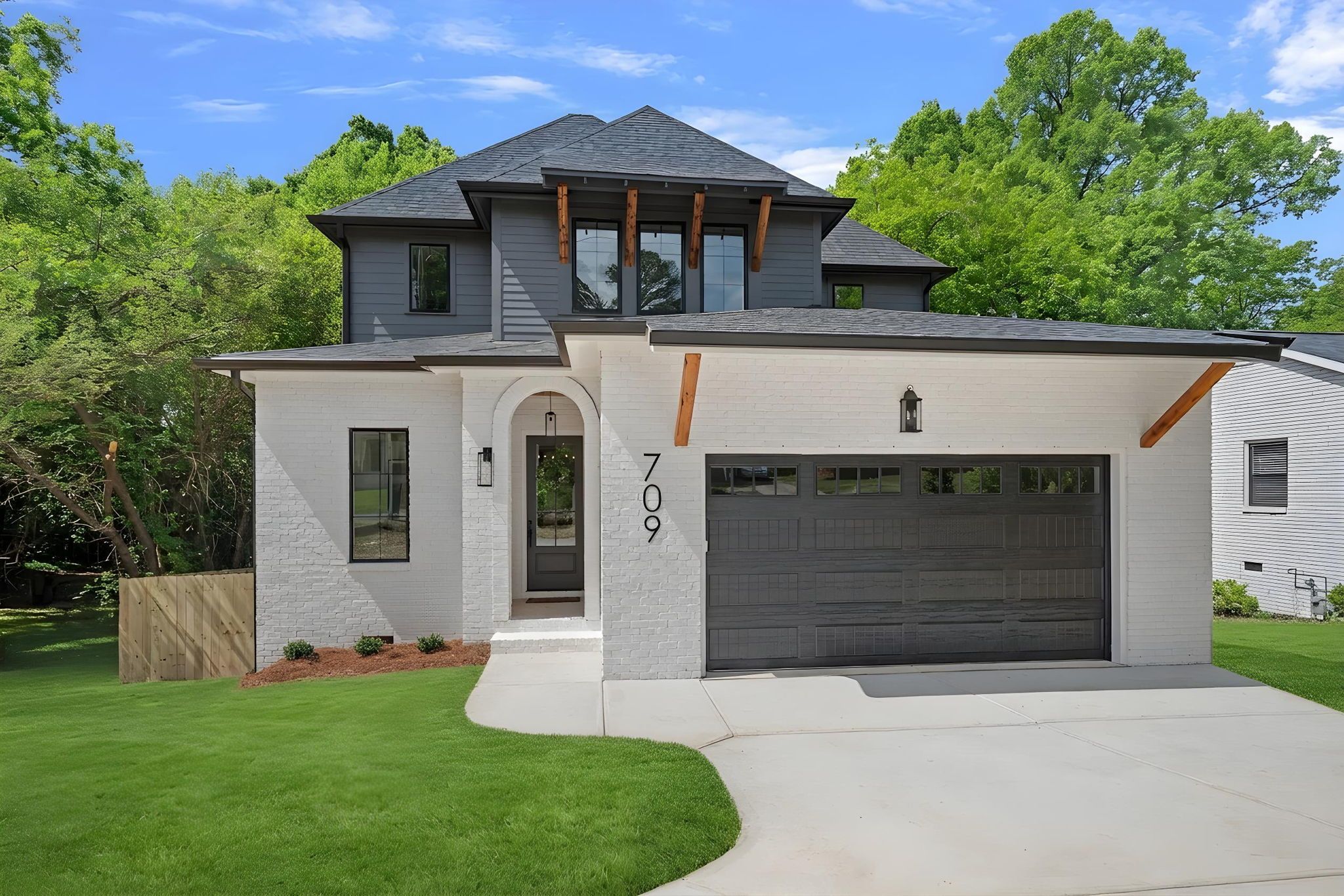 Modern two-story house with white brick base and dark gray siding, large front windows, an arched entry, exposed wooden beams under the eaves, a gray garage door, and green lawn with trees.