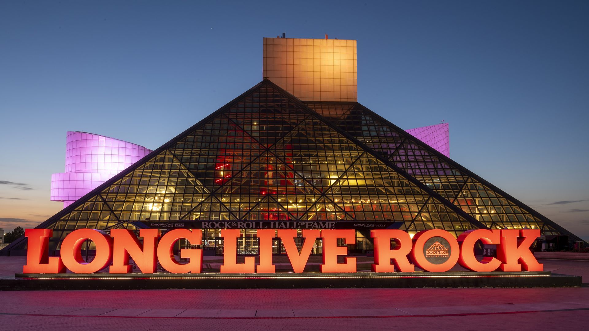 Twilight view of the Rock and Roll Hall of Fame’s glass pyramid, pink side lights, and bold red letters in front reading "LONG LIVE ROCK" against a blue-to-orange sky.