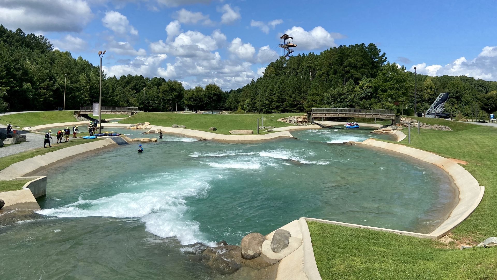A view of the rapids at the Whitewater Center with a zipeline tower off in the distance sticking out from the trees. The sky is blue with scattered clouds. People walk along the edge of the water. 