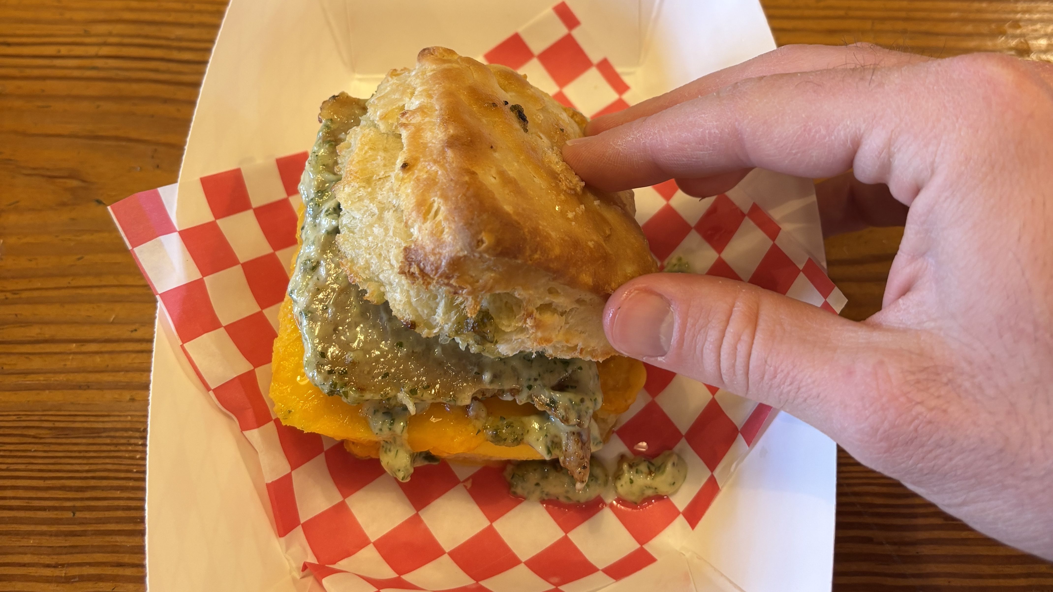 Hand lifting the top of a biscuit sandwich showing melted cheese and creamy green sauce inside, on red and white checkered paper in a white tray, set on a wooden table at Take Care by Oleo.