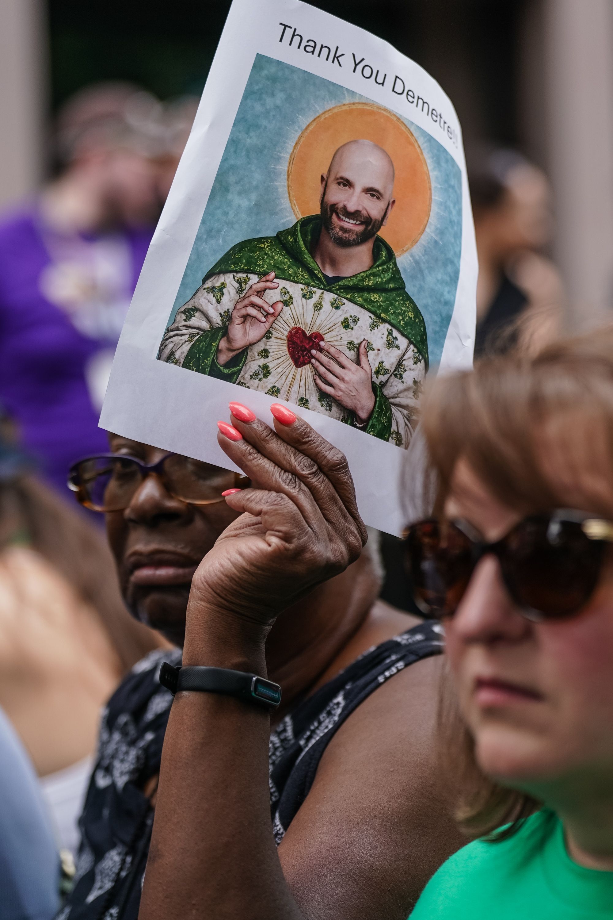 A person with coral pink nails holds a paper sign showing a smiling bald man with a beard, dressed in green and white religious-style robes with a golden halo and a heart symbol, captioned "Thank You Demetr".