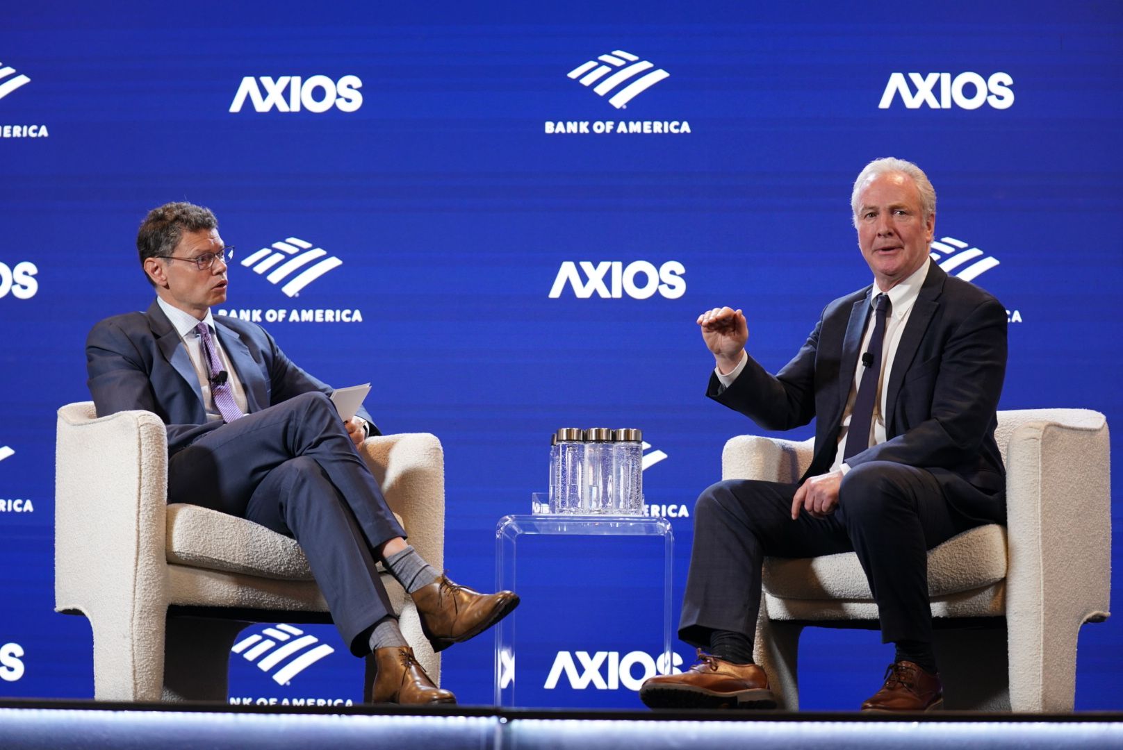 Two men in dark suits sit in cream armchairs on a blue Axios stage, a clear acrylic table with water bottles between them; one man on left looks at papers, the other on right gestures with a fist.