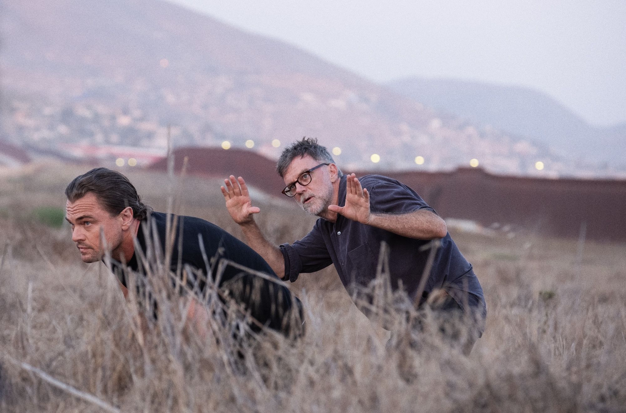 Director-writer-producer Paul Thomas Anderson and Leonardo DiCaprio crouch in dry grass; one wears black and leans forward, the other in glasses and dark shirt gestures with raised hands. Hills and blurred lights in the background.