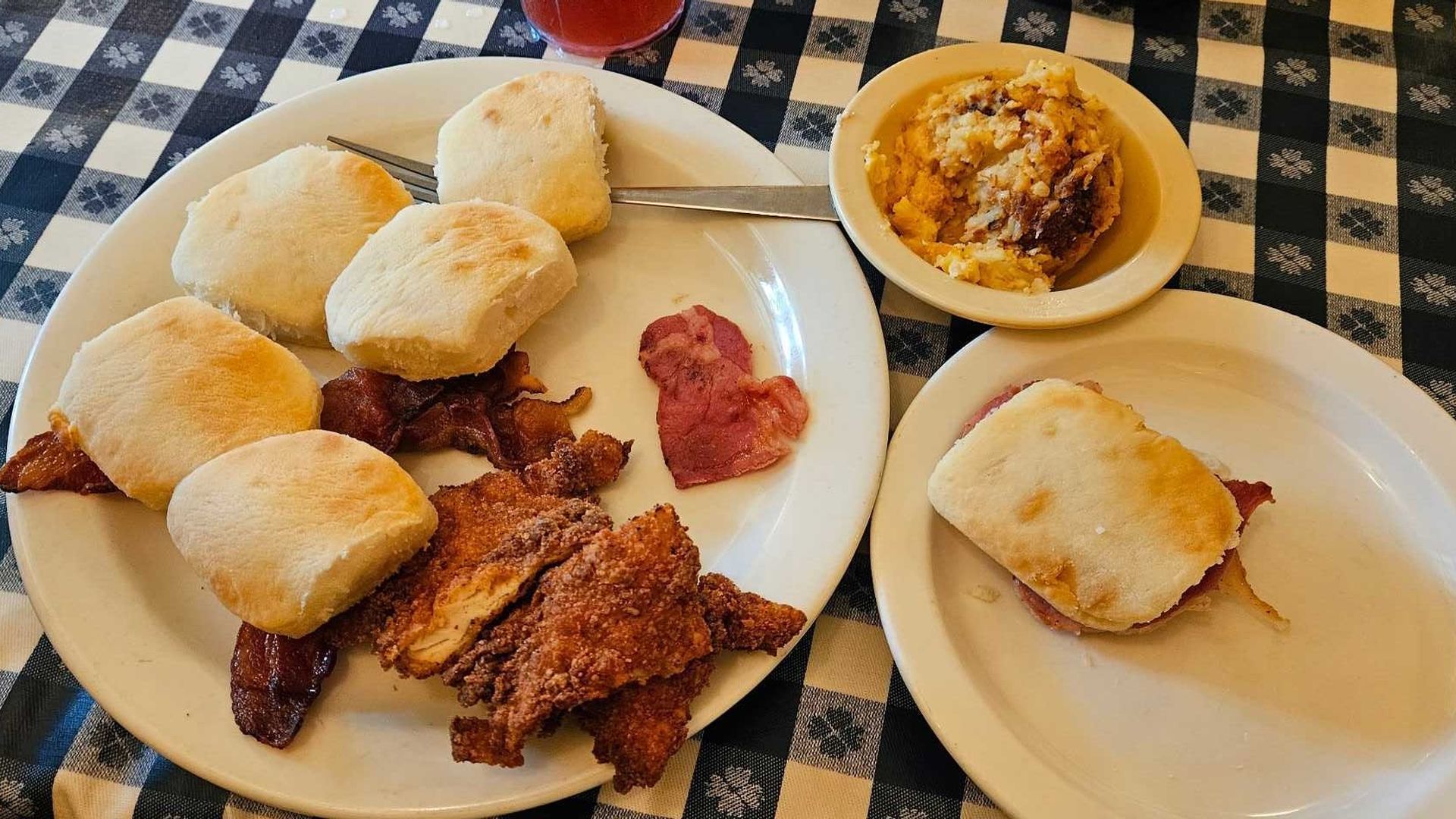 Top-down view of a table with a blue-and-white checkered cloth, showing three plates of food: fluffy biscuits, fried chicken, ham slices, bacon, and a beige bowl with a baked casserole.