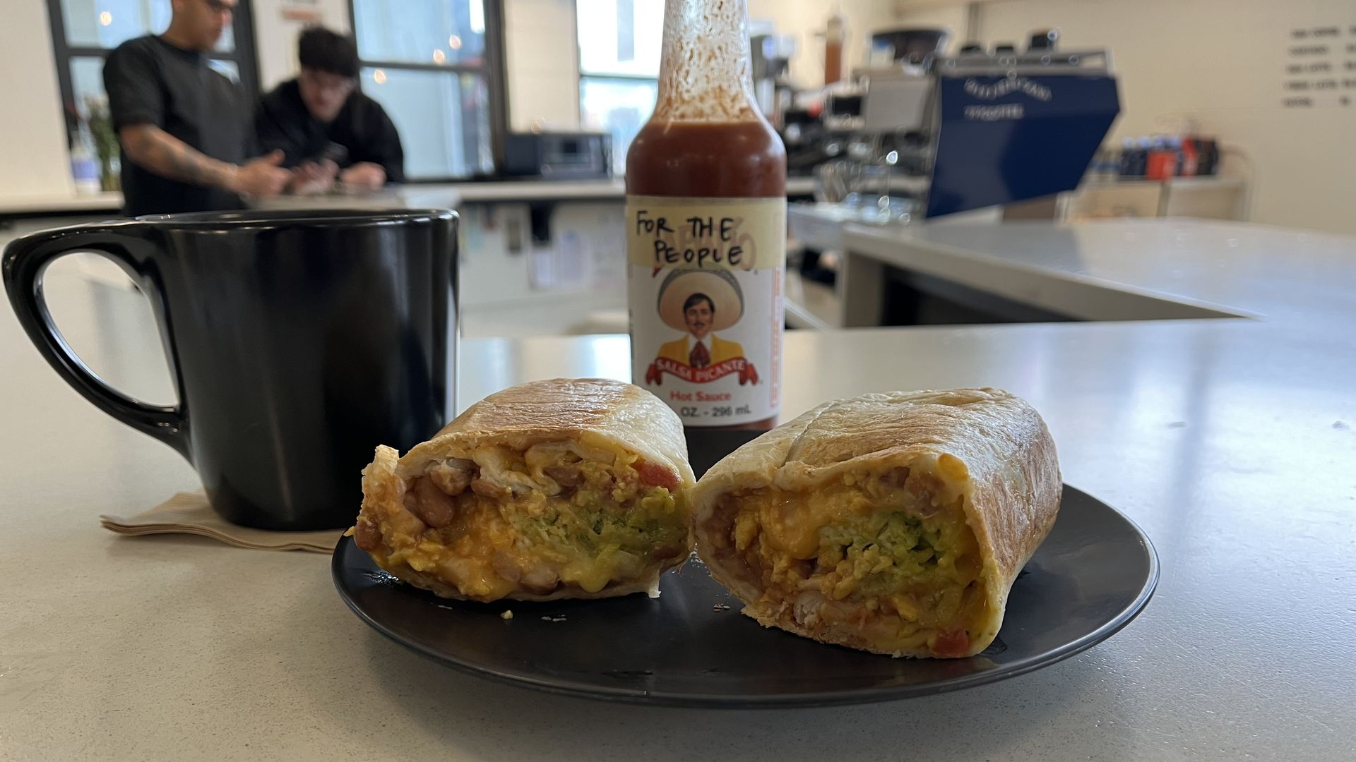 The burrito sits on a plate with coffee and hot sauce next to it.
