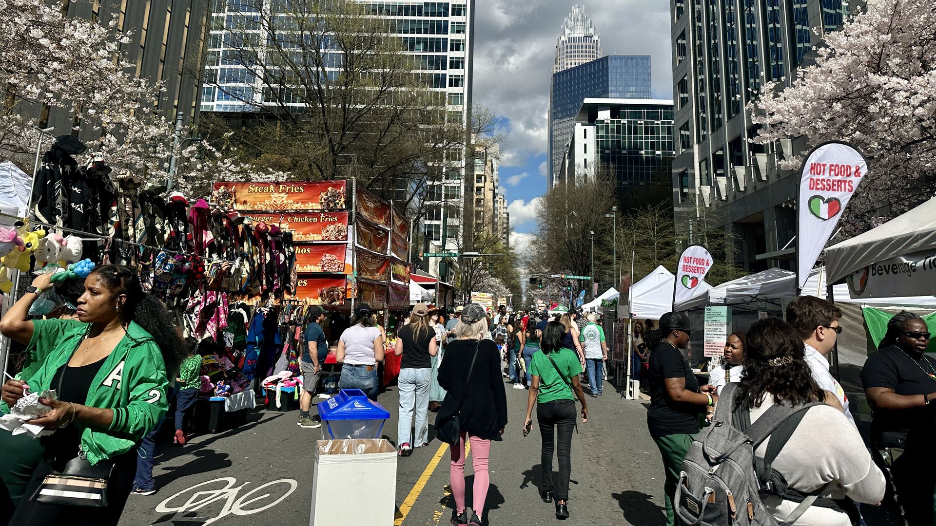 City street festival with vendor stalls along a tree-lined avenue, pink blossoms, and tall glass skyscrapers. Crowds walk past an orange "Steak Fries" truck and "Hot Food & Desserts" banners.