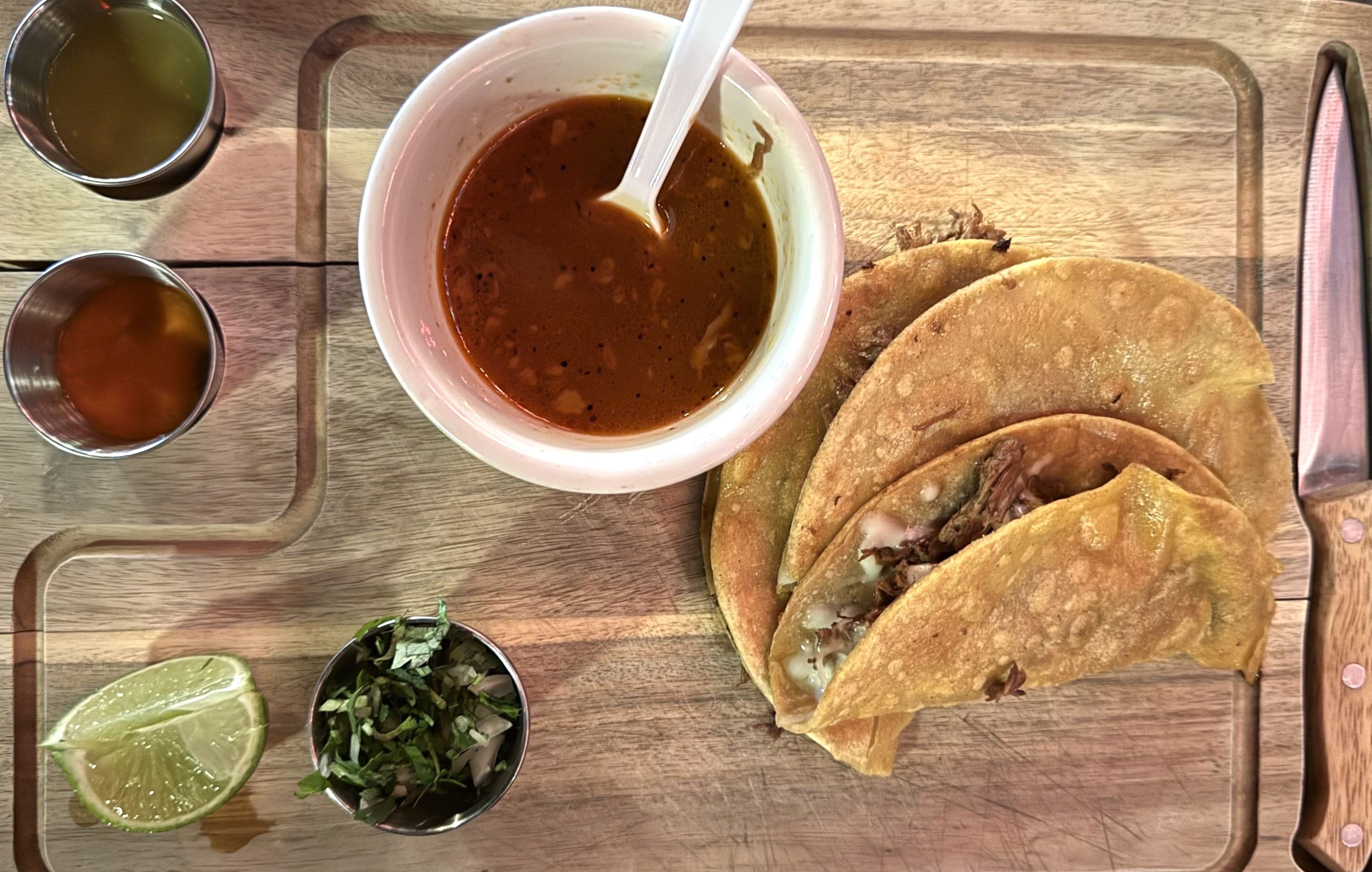 Three birria tacos with melted cheese are served on a wooden board alongside a bowl of consommé, lime wedges, chopped onions and cilantro, and two small cups of salsa. A steak knife rests on the side.