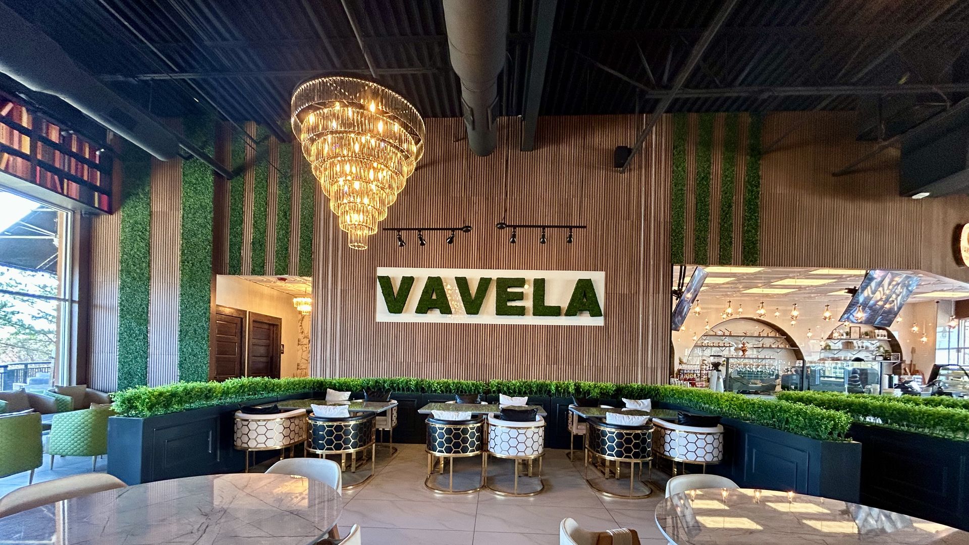 Bright cafe interior with marble tables, beige and black chairs, green plants lining the seating area, a large gold chandelier, and the word "VAVELA" displayed in green on a wood-paneled wall.