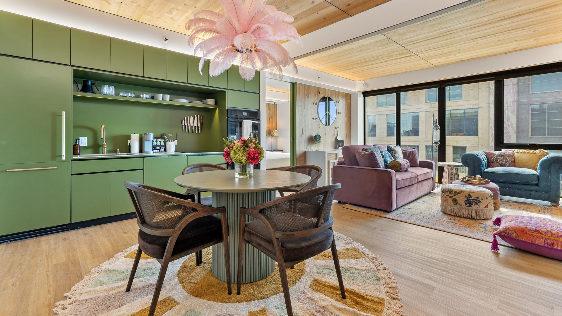 Modern open living area with green kitchen cabinets, round dining table with dark chairs on a colorful rug, pink feather chandelier, purple and blue sofas, large windows, and wooden ceiling.