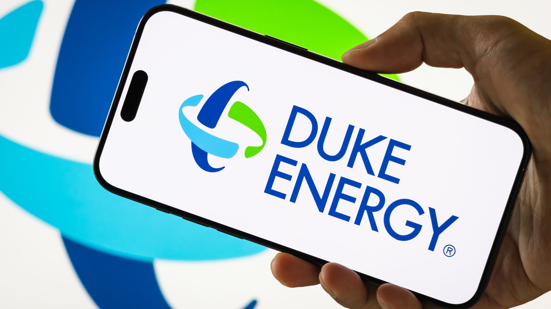Hand holding a smartphone displaying the Duke Energy logo with blue and green elements on a white background, with a larger blurred logo behind it.
