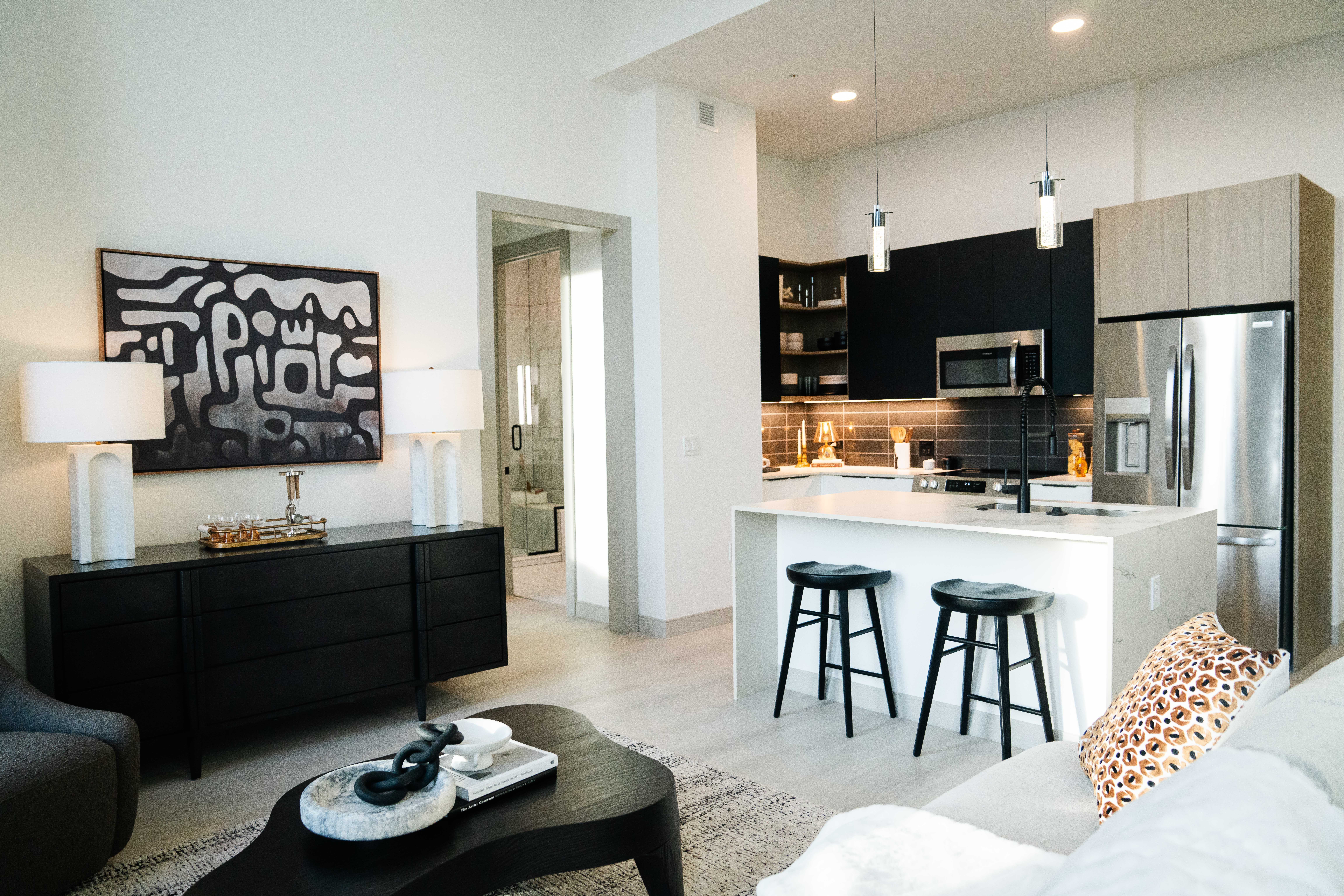 Modern open-concept living room and kitchen with black and white decor, abstract black and white wall art, black furniture, two white lamps, kitchen island with two black stools, and stainless steel fridge.
