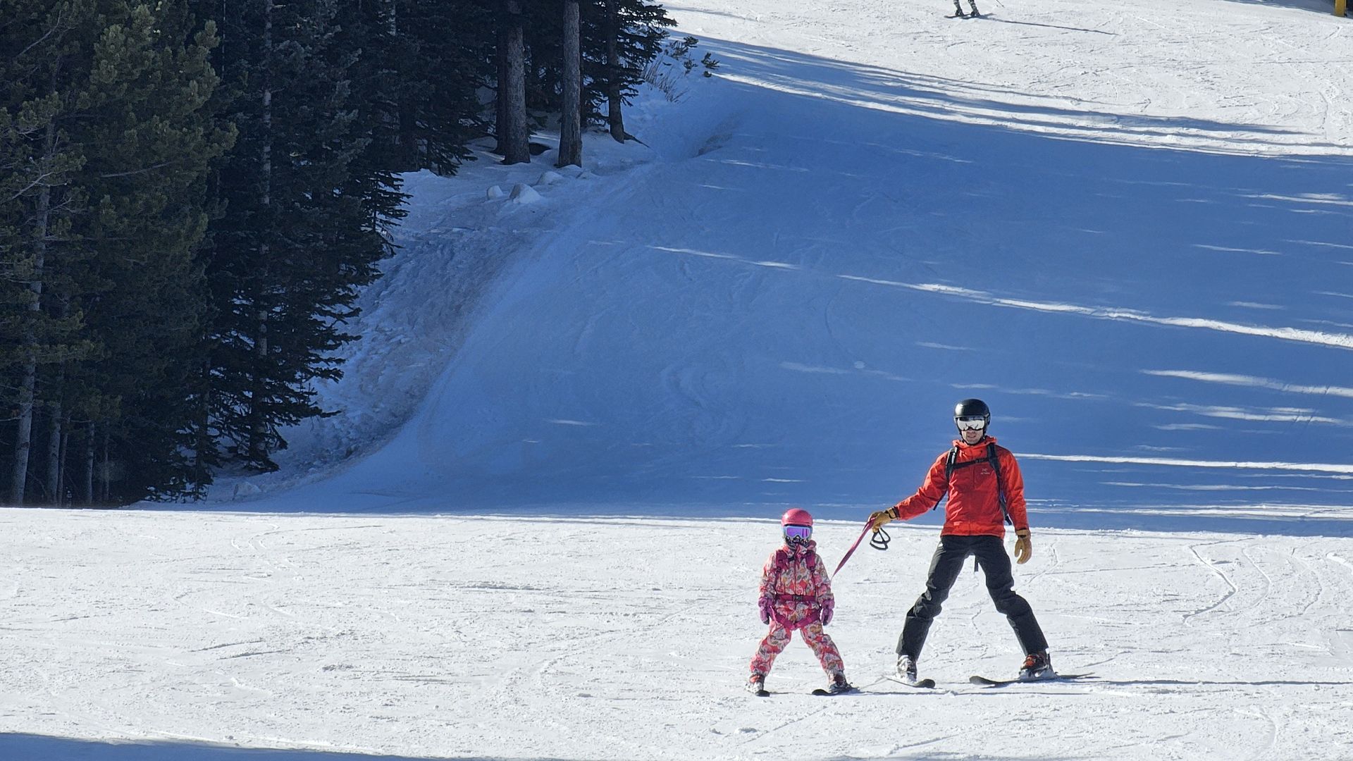 A man in a red jacket skis with a child in a pink snowsuit on a snowy slope, with a pine forest to the left and a distant skier in the background.