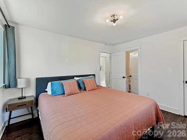 Minimalist bedroom with white walls, dark wood floors, large bed with pink quilt and teal and pink pillows, dark headboard, small bedside table with lamp, and two white doors.