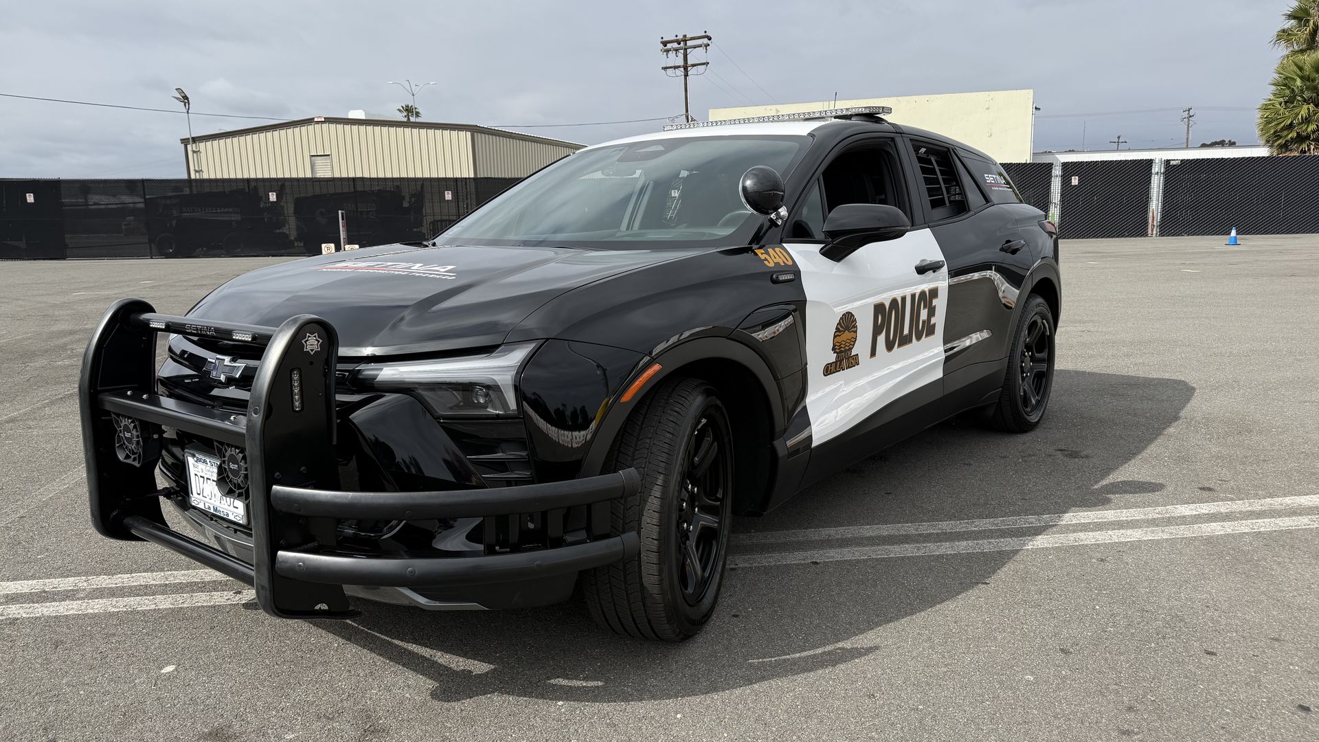 Front-left view of a Chevy Police Pursuit Vehicle SUV in black-and-white livery, with a push bar, roof light bar, and POLICE on the side, parked in a lot with a chain-link fence and cloudy sky.