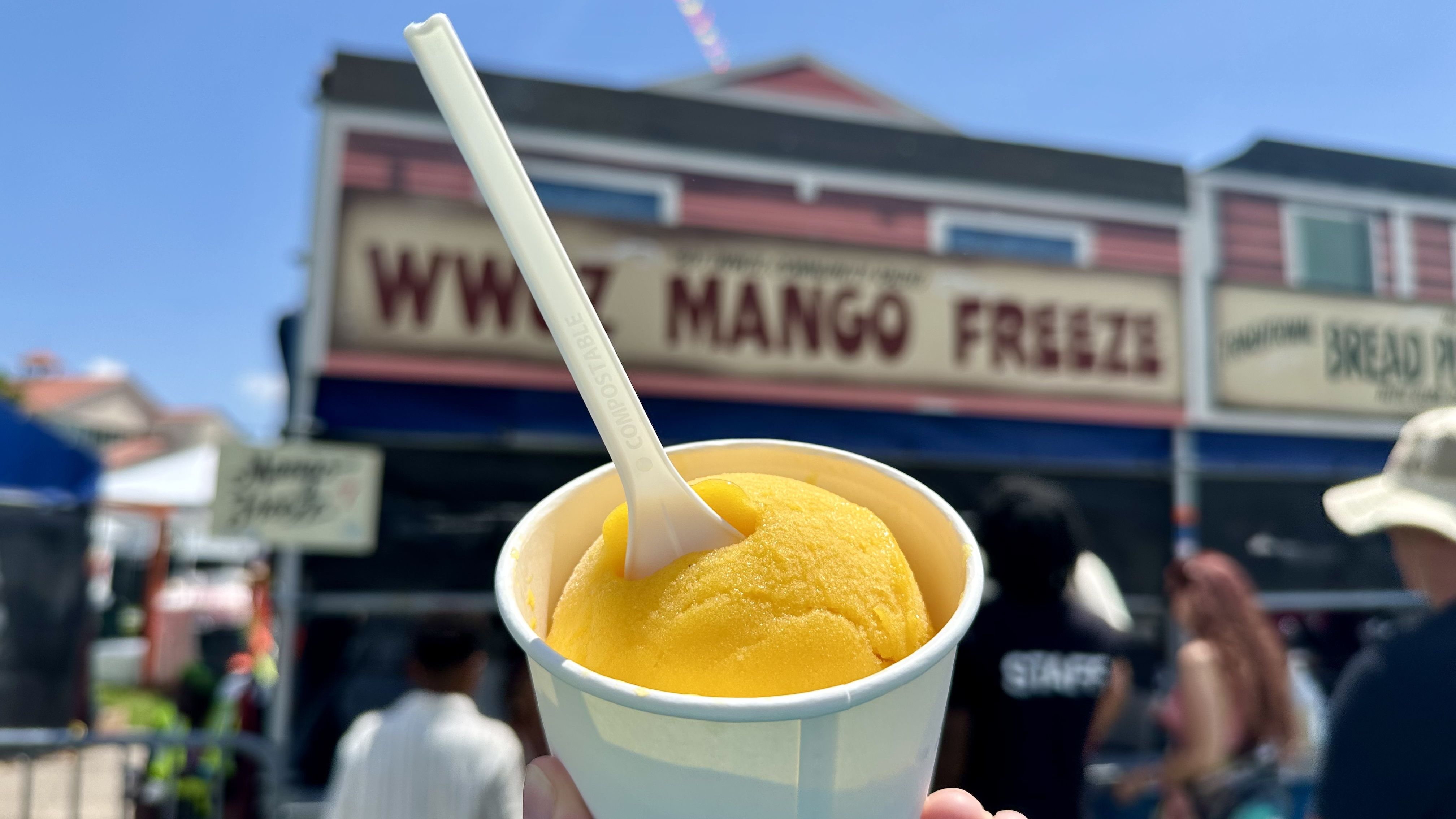 Close-up of bright yellow mango ice cream in a white cup with a compostable spoon, held up in front of a storefront sign reading "MANGO FREEZE" at a sunny outdoor market.