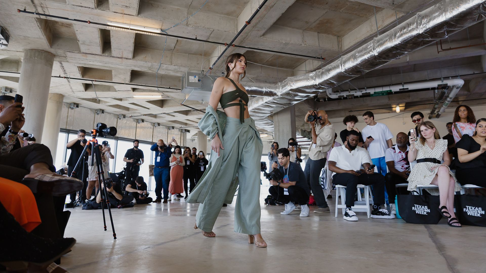 Fashion model in a sage-green outfit walks a runway inside an unfinished concrete space; overhead ducts loom as photographers and an audience watch from both sides.