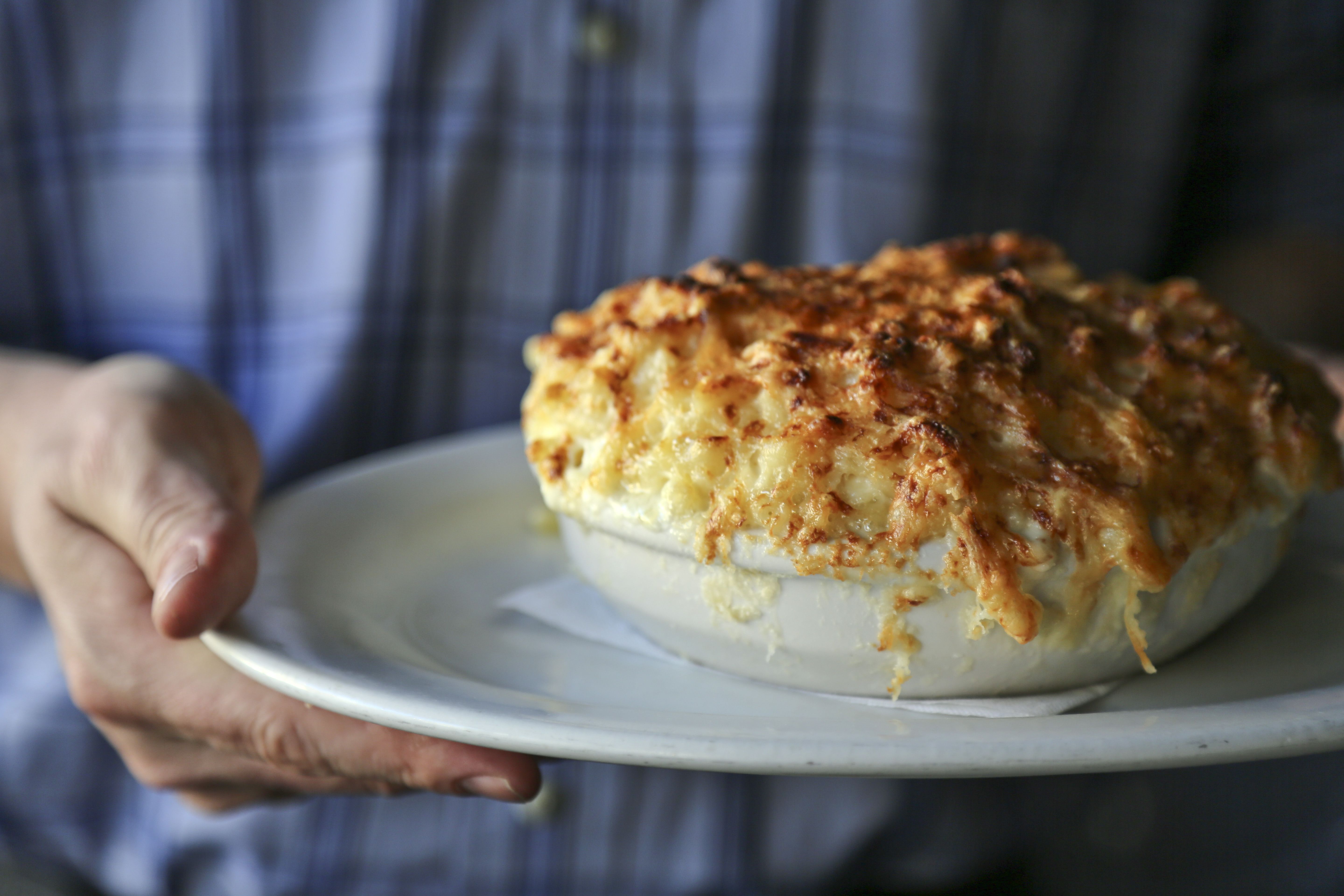 Close-up of a baked cheesy dish with a golden-brown crust on a white plate, held by a person in a blue plaid shirt; bubbly interior, blurred background.