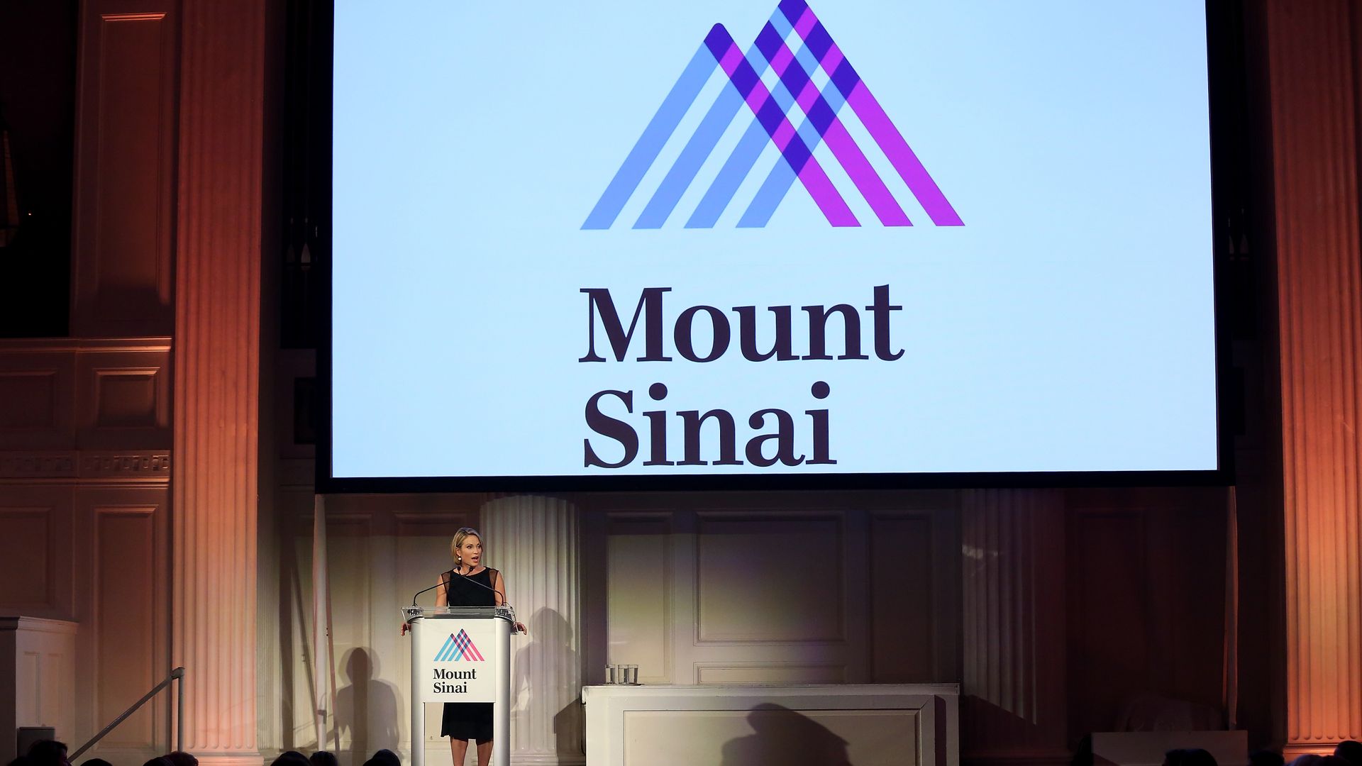 A woman speaks near a large white screen that displays the blue and pink logo for Mount Sinai Health System.